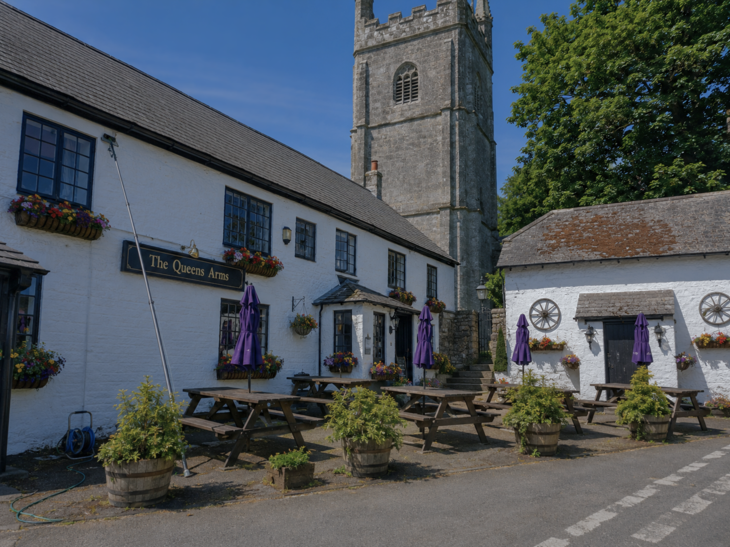 Outdoor patio of The Queens Arms pub with wooden picnic tables, purple umbrellas, and flower baskets on white walls. A tall stone church steeple is visible in the background, along with green trees and a clear blue sky.