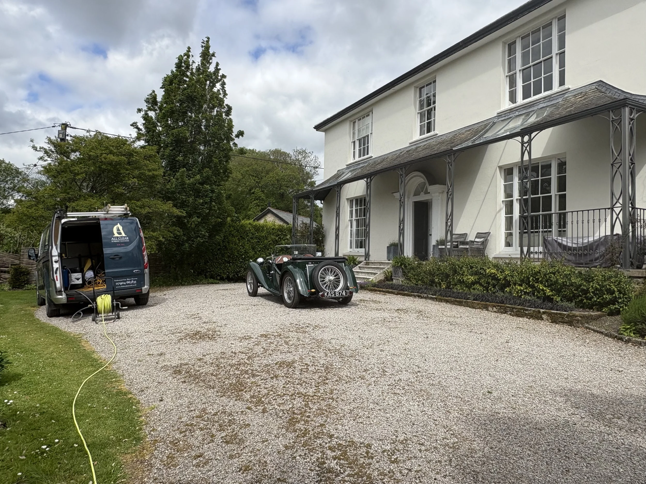 Front yard of a white house with a porch, two black vintage cars parked on a gravel driveway, a utility van with equipment, green trees, cloudy sky.