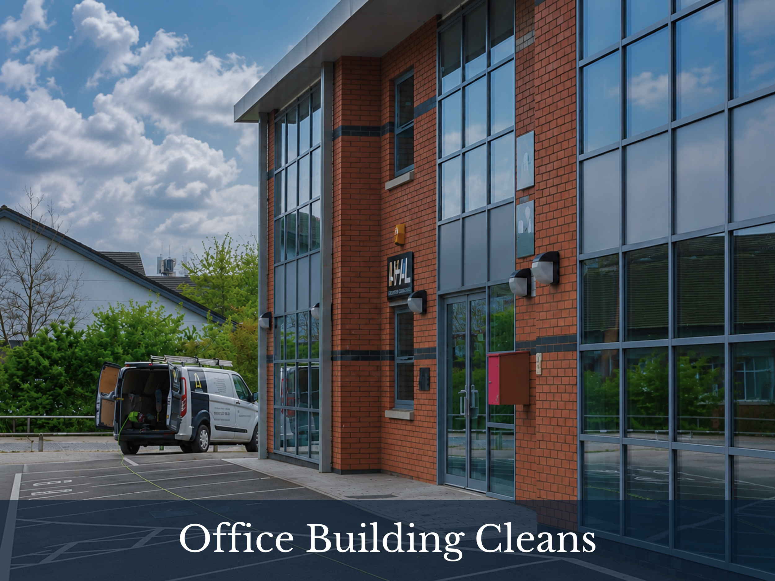 The image shows a modern office building with large glass windows and red brick walls. A van is parked in the parking lot in front of the building, connected to a power source for cleaning or maintenance. The sky is partly cloudy with some patches of blue visible. There are some trees and a house in the background.