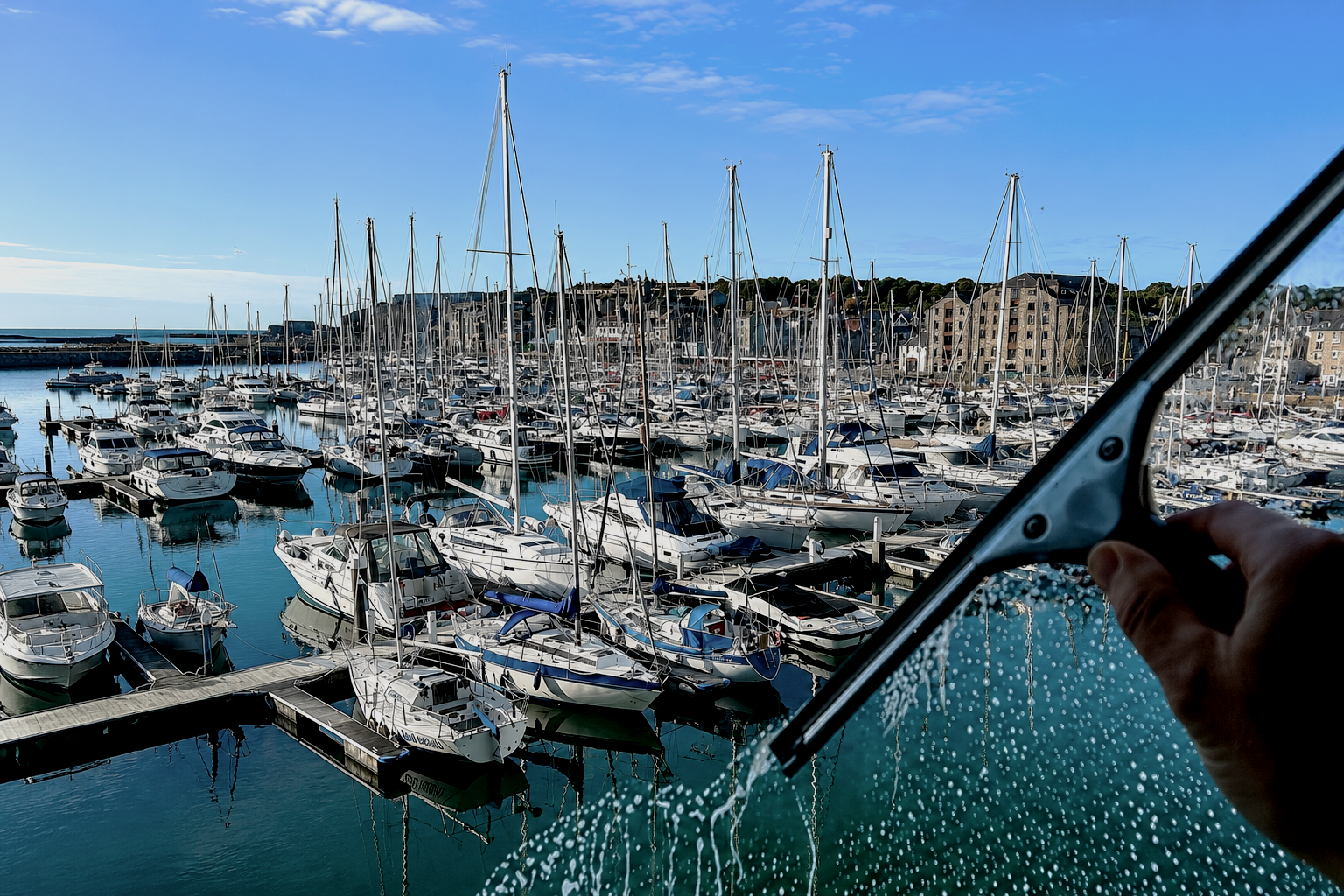 A person's hand holding a windshield wiper while cleaning a marina window overlooking numerous docked boats and yachts with sailboats, with a clear blue sky and some buildings in the background.