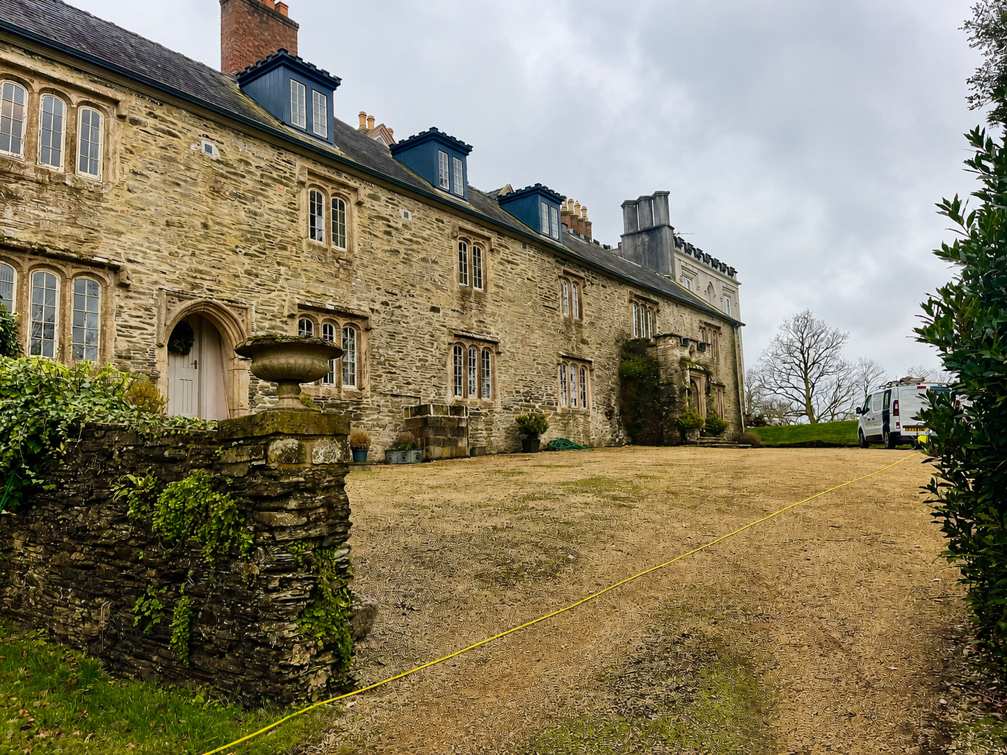 Stone historic mansion with multiple windows and blue dormer roofs, a gravel driveway, and a white van parked to the right, on a cloudy day with leafless trees in the background.