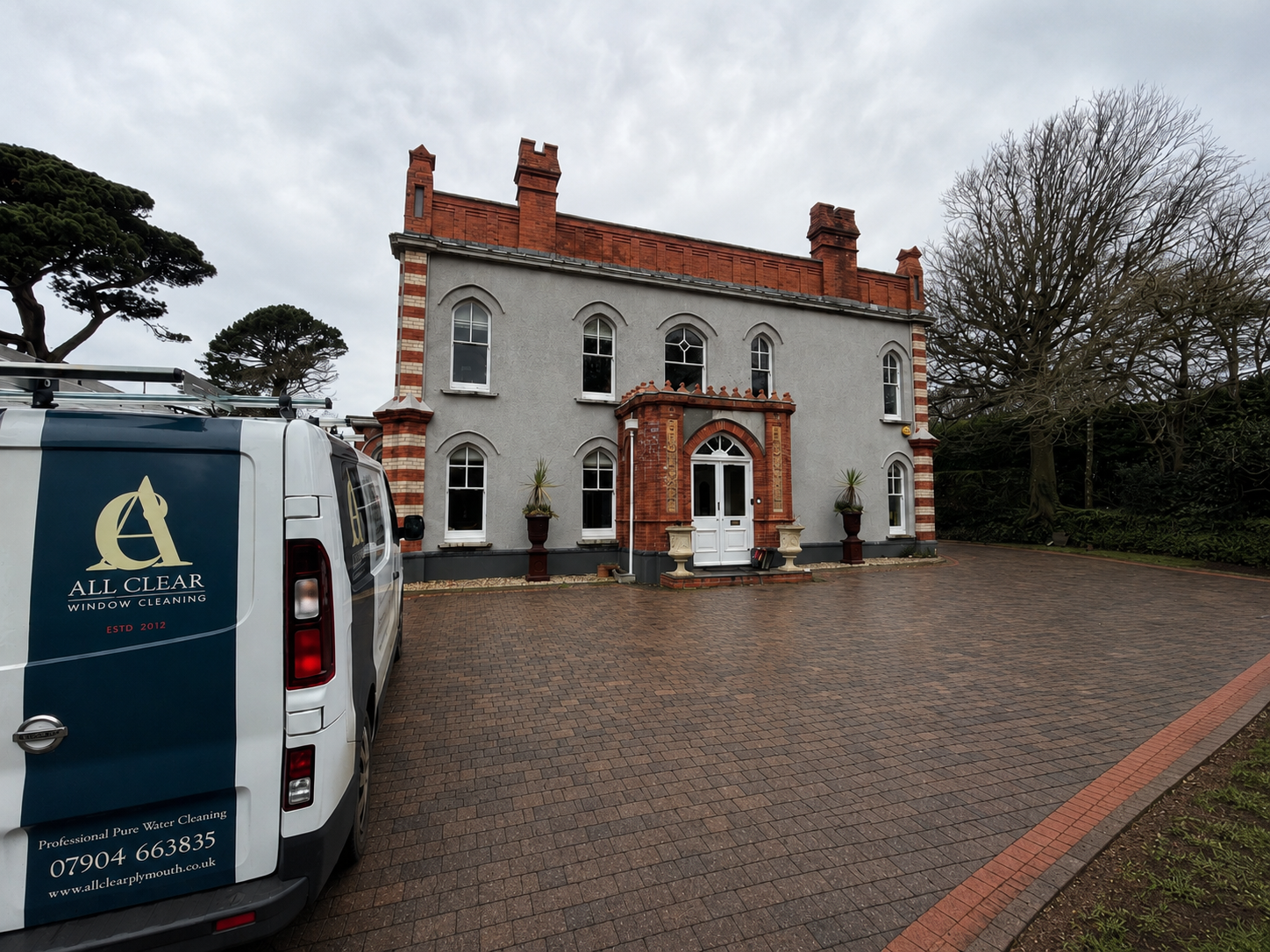 A large, three-story house with a brick and gray exterior, multiple arched windows, and potted plants on either side of the front door. A cleaning service van labeled "All Clear Window Cleaning" is parked in the driveway made of brick pavers. Trees and cloudy sky in the background.