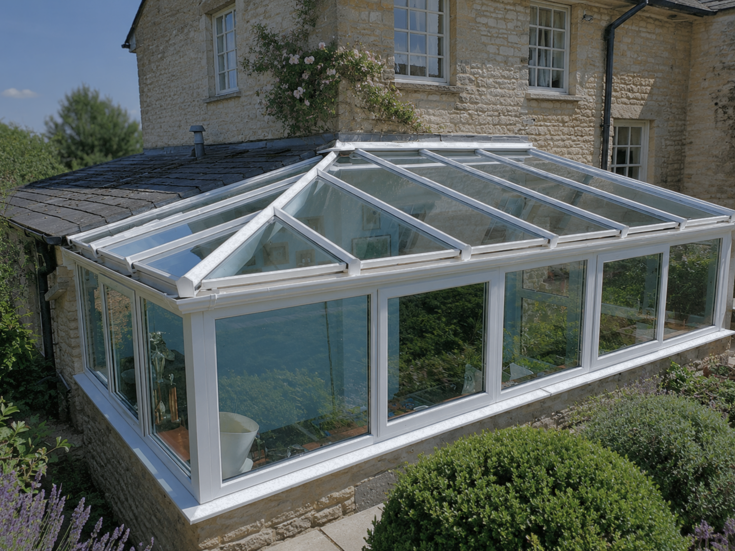 A glass conservatory attached to a stone house, with a sloped roof and white framing, surrounded by greenery and flowering plants.