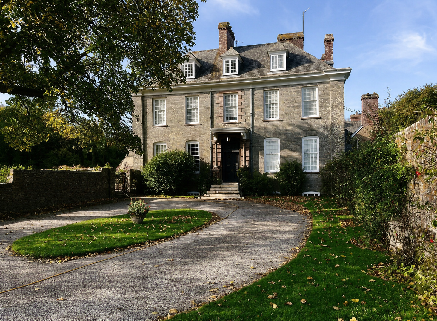 A large, historic stone house with a slate roof and multiple chimneys, surrounded by green lawn and trees, with a curved driveway leading to the front entrance.