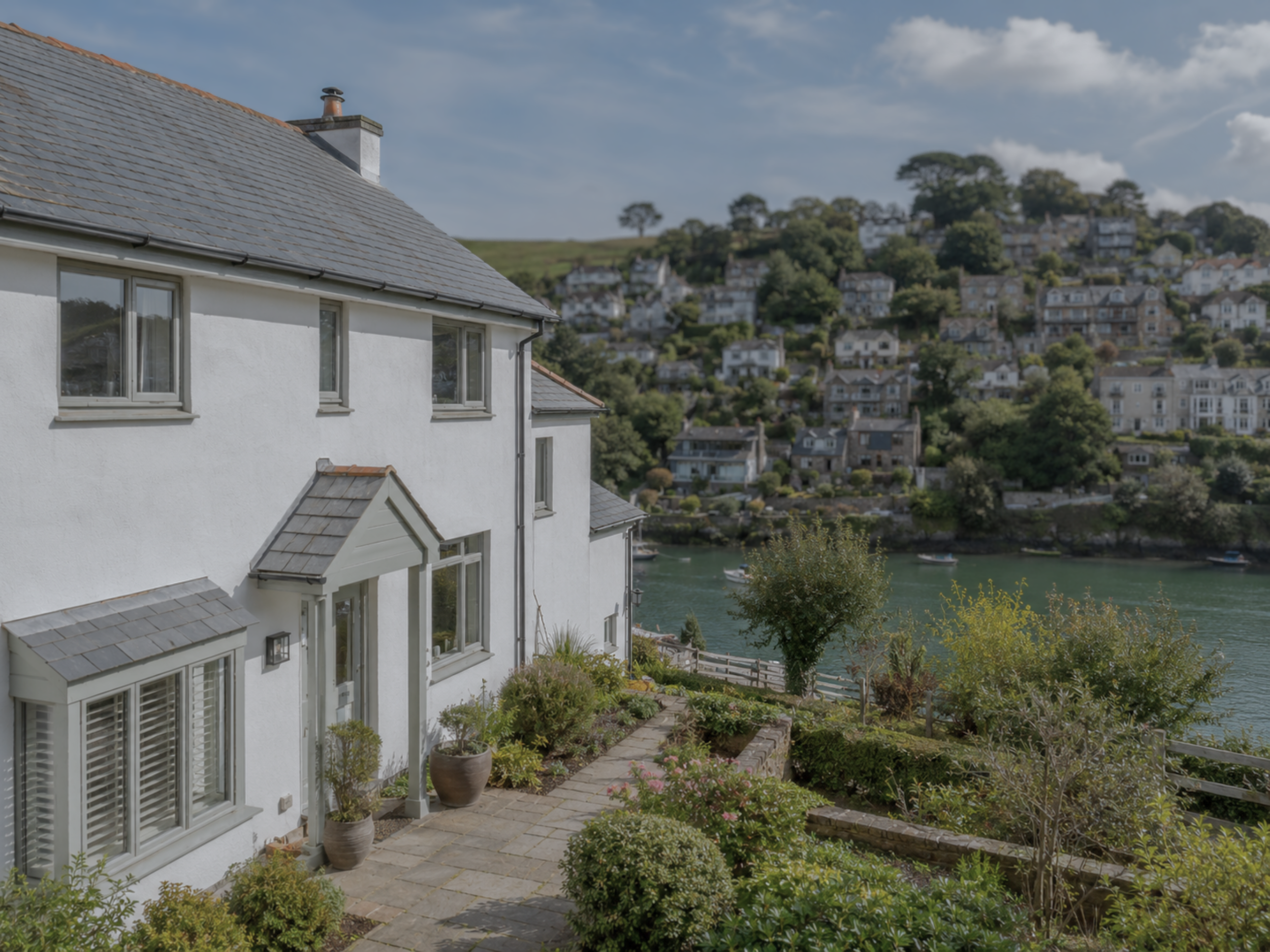 White house with gray roof, potted plants, stone patio, overlooking a river with boats and a hillside filled with houses and trees under a partly cloudy sky.