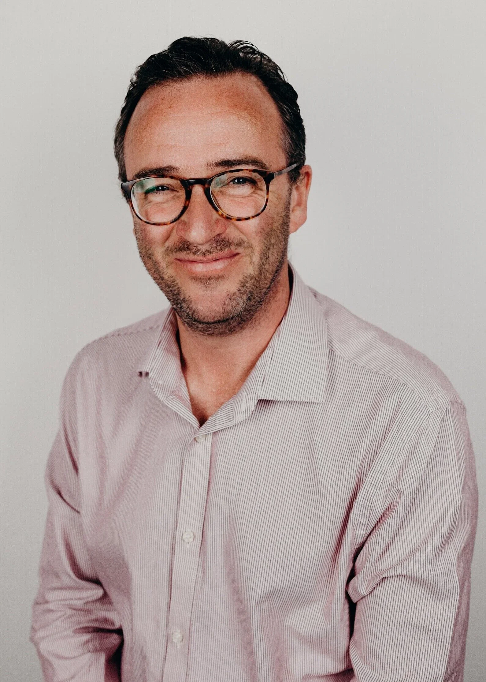 Headshot of a smiling man with dark hair, glasses, a beard, wearing a light pink button-up shirt, against a plain white background.