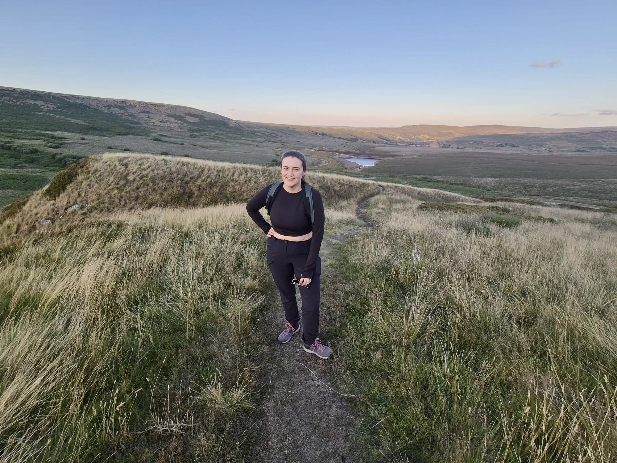 A woman standing on a trail in a grassy hilly landscape during sunset, with a lake in the distance.