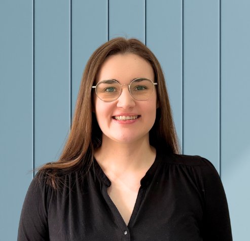 A woman with long brown hair wearing glasses and a black blouse stands in front of a blue wooden paneled wall, smiling at the camera.