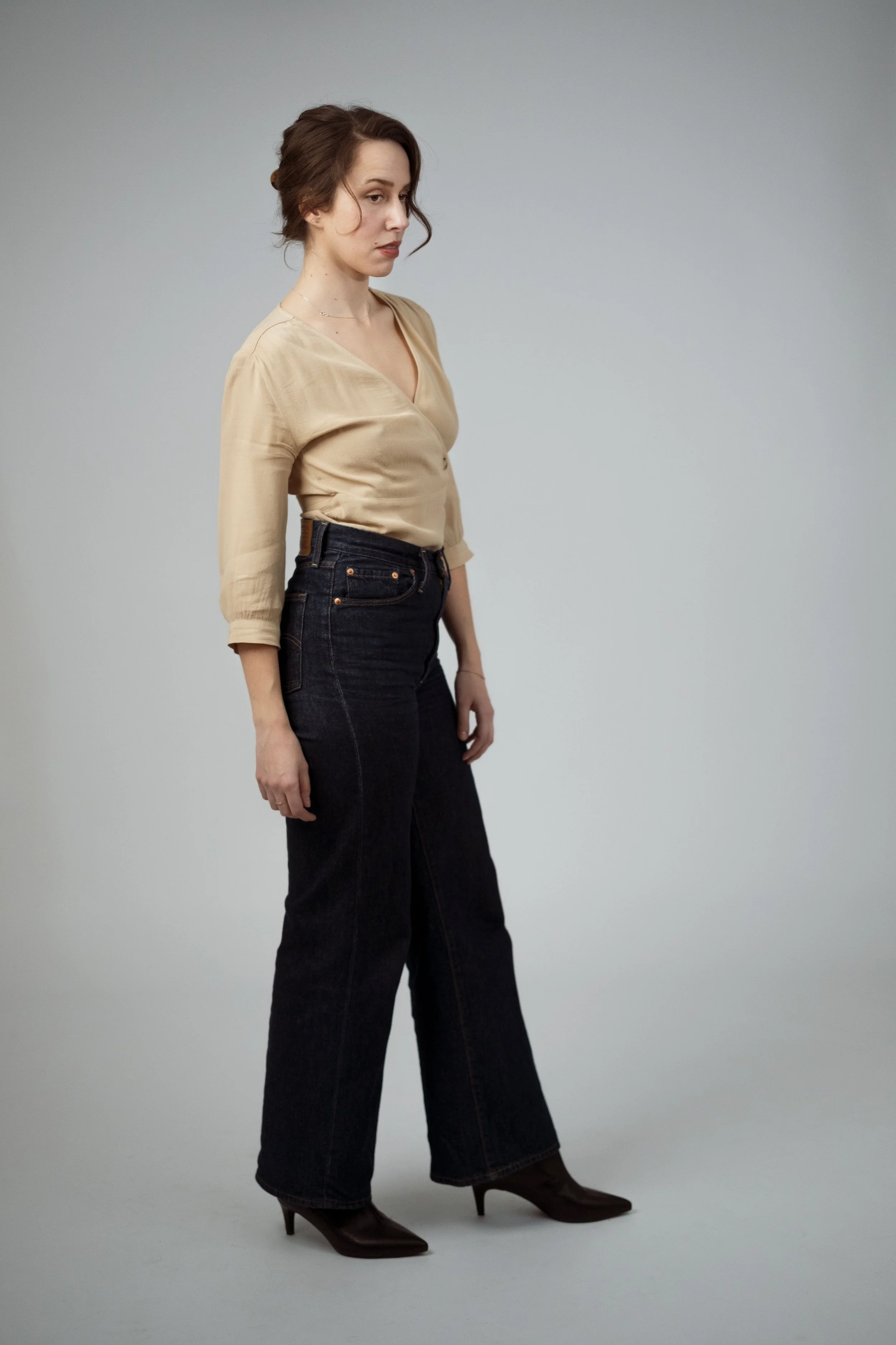 En ung kvinna med brunt lockigt hår, bär en beige blus, svarta jeans och höga stövlar, står mot en grå bakgrund.