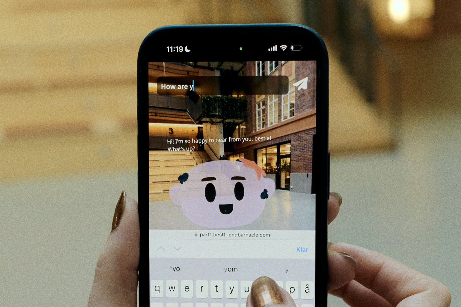 En hand håller en smartphone som visar en konversation med en animerad molnfigur med ansikte. Bakom syns en byggnad med trappor och fönster.