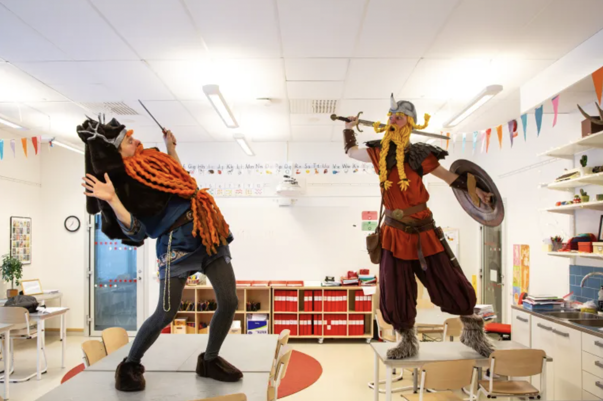 Två personer i vikingdräkter leker i ett klassrum, en är utklädd till en viking och håller ett svärd, den andra till en annan viking med hornad hjälm och sköld.