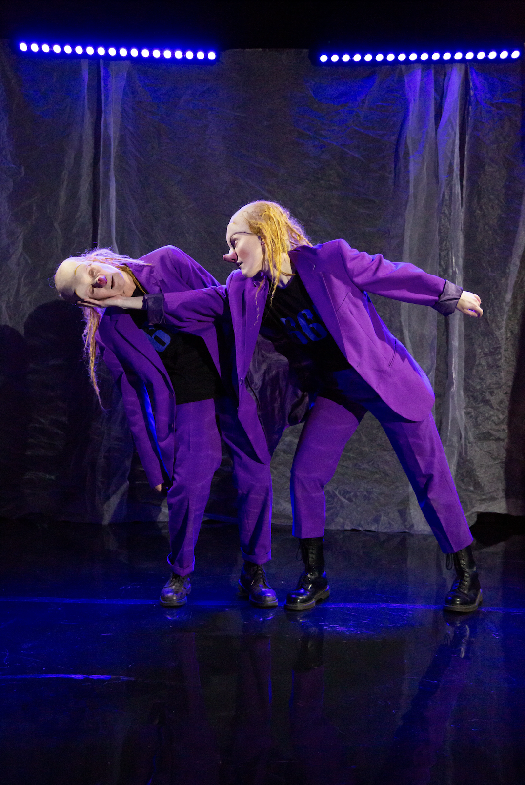 Två personer i stilrena, mörka kläder och lila kostymer som uppträder på scen under en nattlig ljusshow.