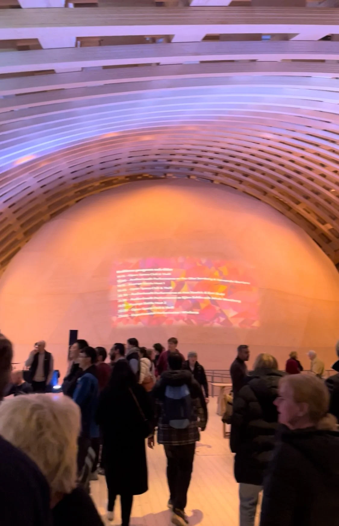 Människor samlade inomhus i en stor byggnad med en stor kurvad vägg. Det finns en ljus projicerad bild på väggen och ett antal personer står och pratar.