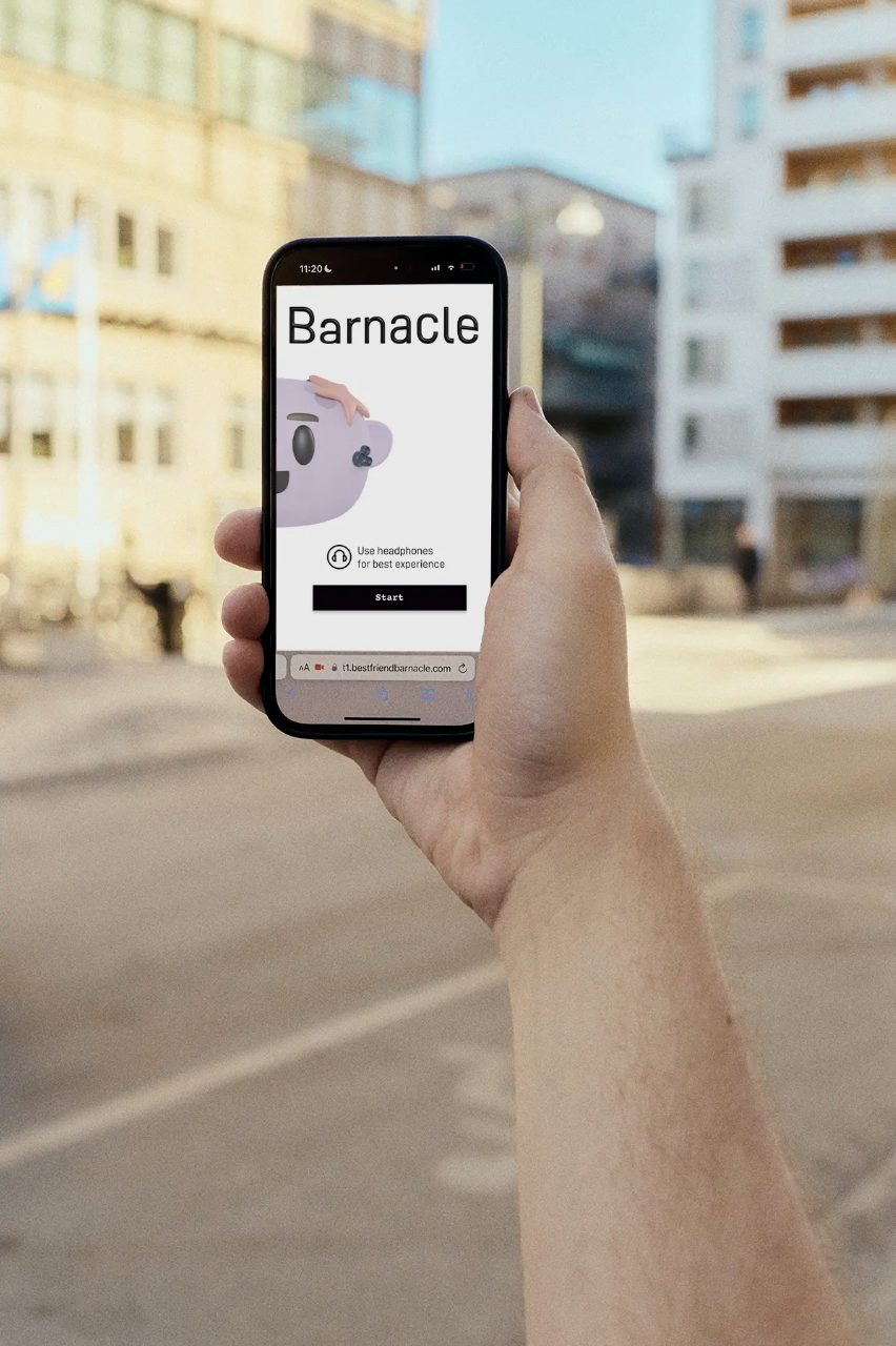 En person håller en mobiltelefon som visar en app för Barnacle, med ett tecknat valrossansikte på skärmen, i ett stadsmiljö.