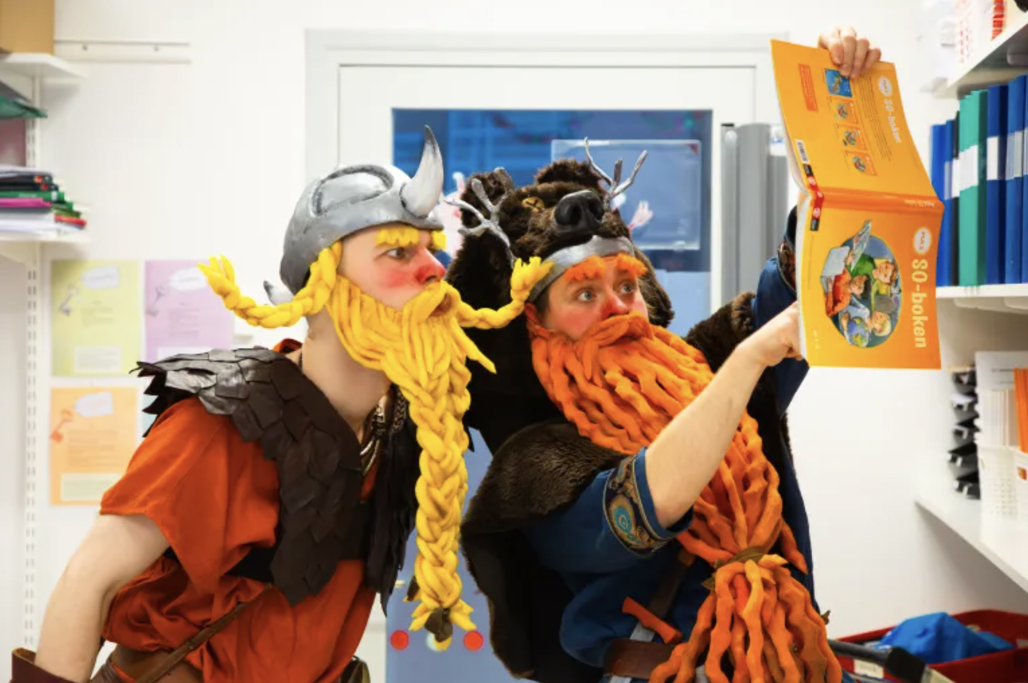 Två personer klädda som vikingar står i ett klassrum och tittar på en bok, alla har färgstarka skägg av garn och bär hjälmar och andra vikingainspirerade kostymer.