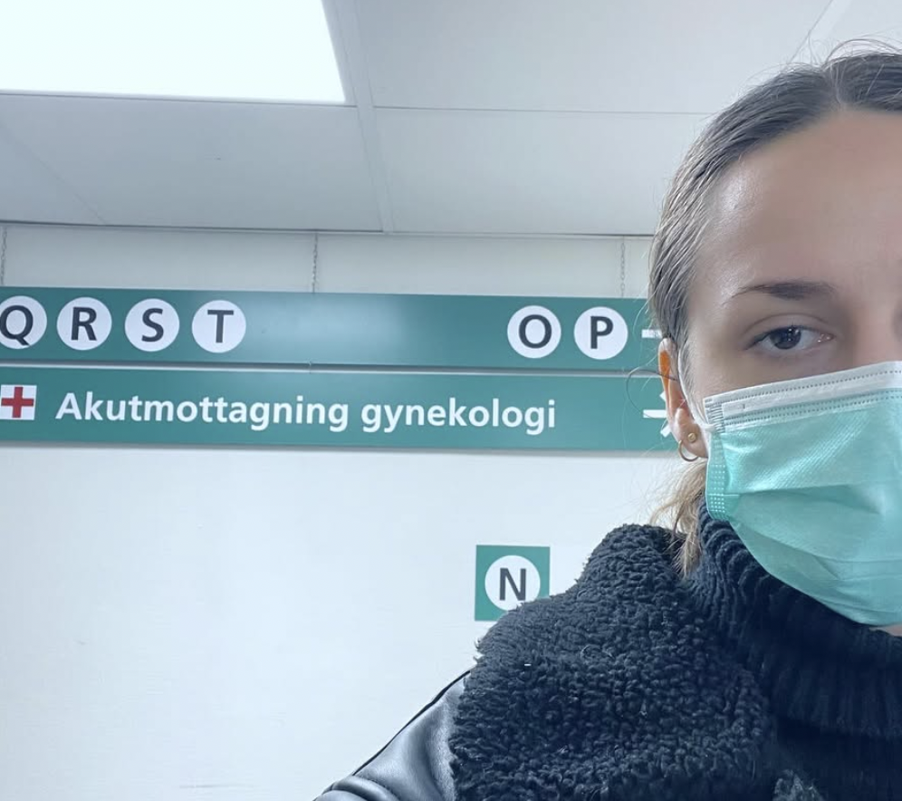 En person med munskydd på en sjukhusavdelning för gynekologi, uppsatt vägskylt för akutmottagning.