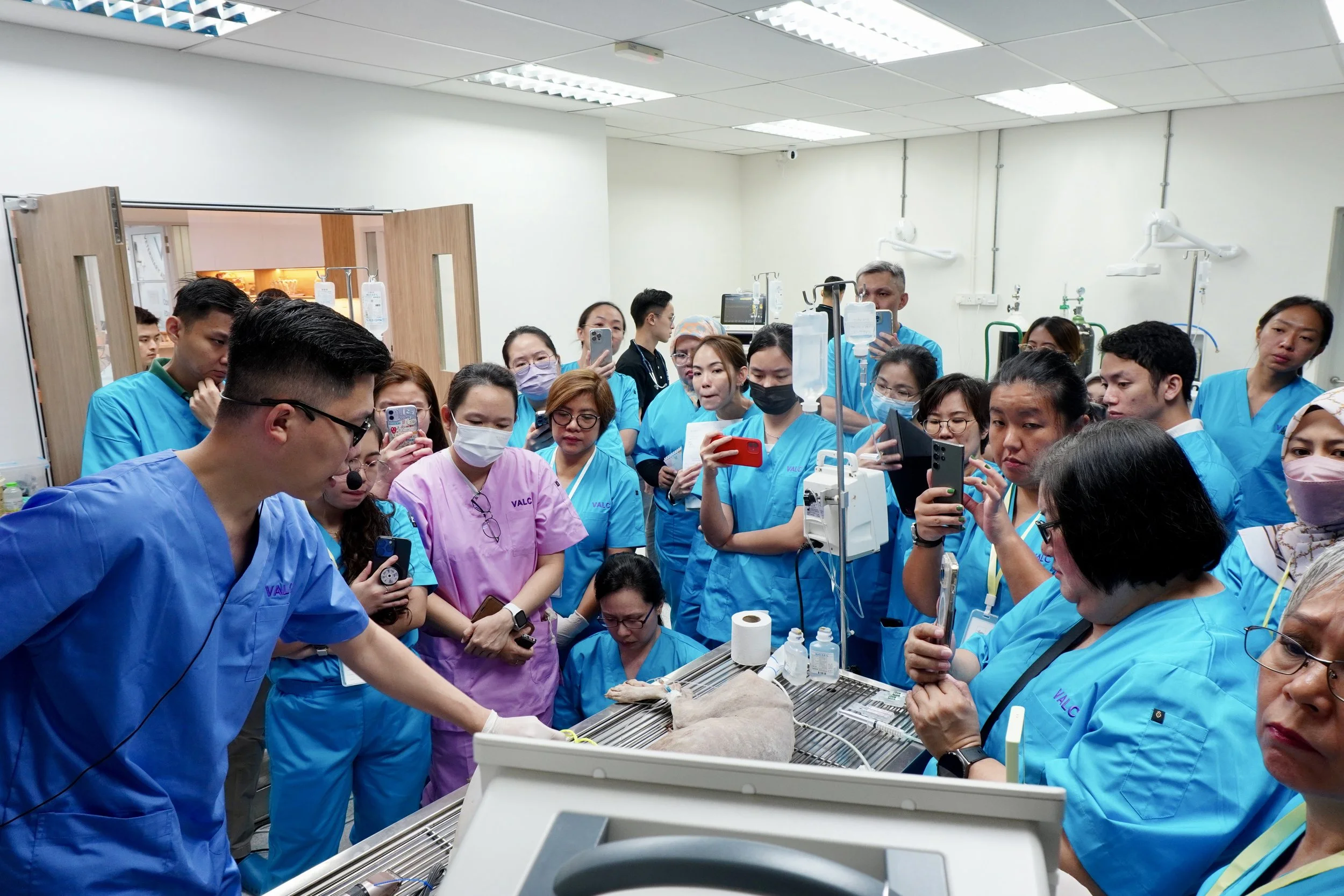 Group of VALC students in scrubs gathered around a hospital bed, observing a medical procedure in a clinical setting.