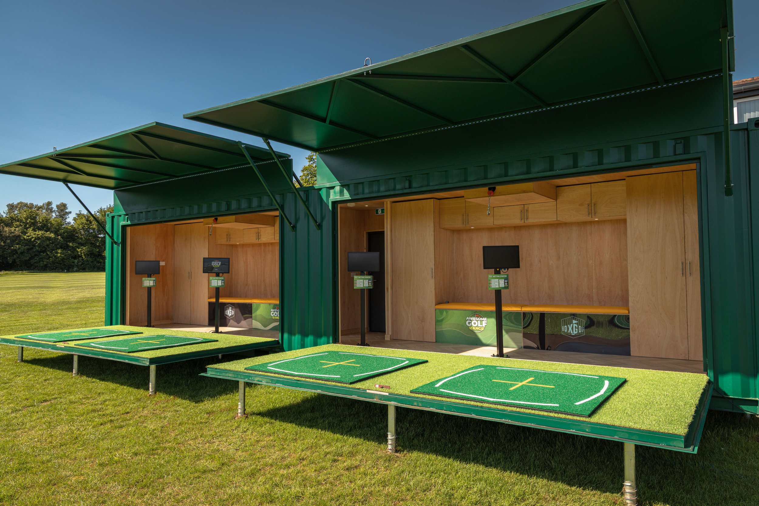 A portable golf driving range made of shipping containers with open lids, featuring small golf mats and monitors, set up on a grassy field under a clear blue sky.