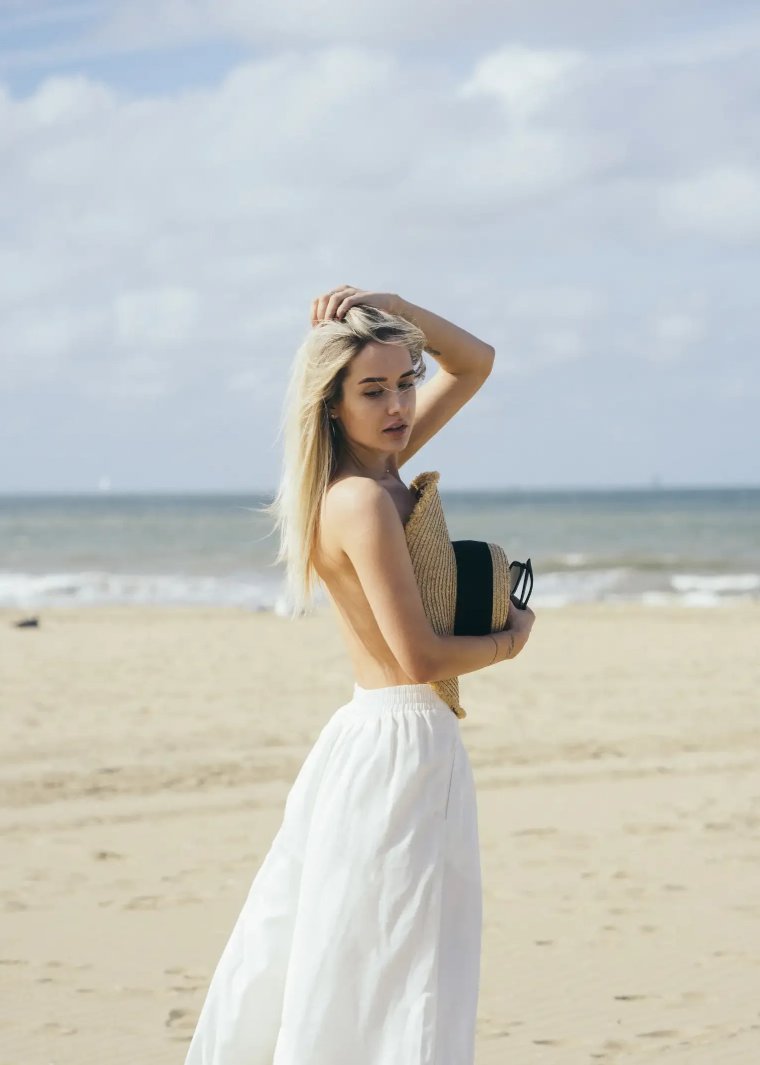 A woman in a beige top and white skirt standing on a sandy beach with the ocean in the background, holding her hair back with one hand against a partly cloudy sky.