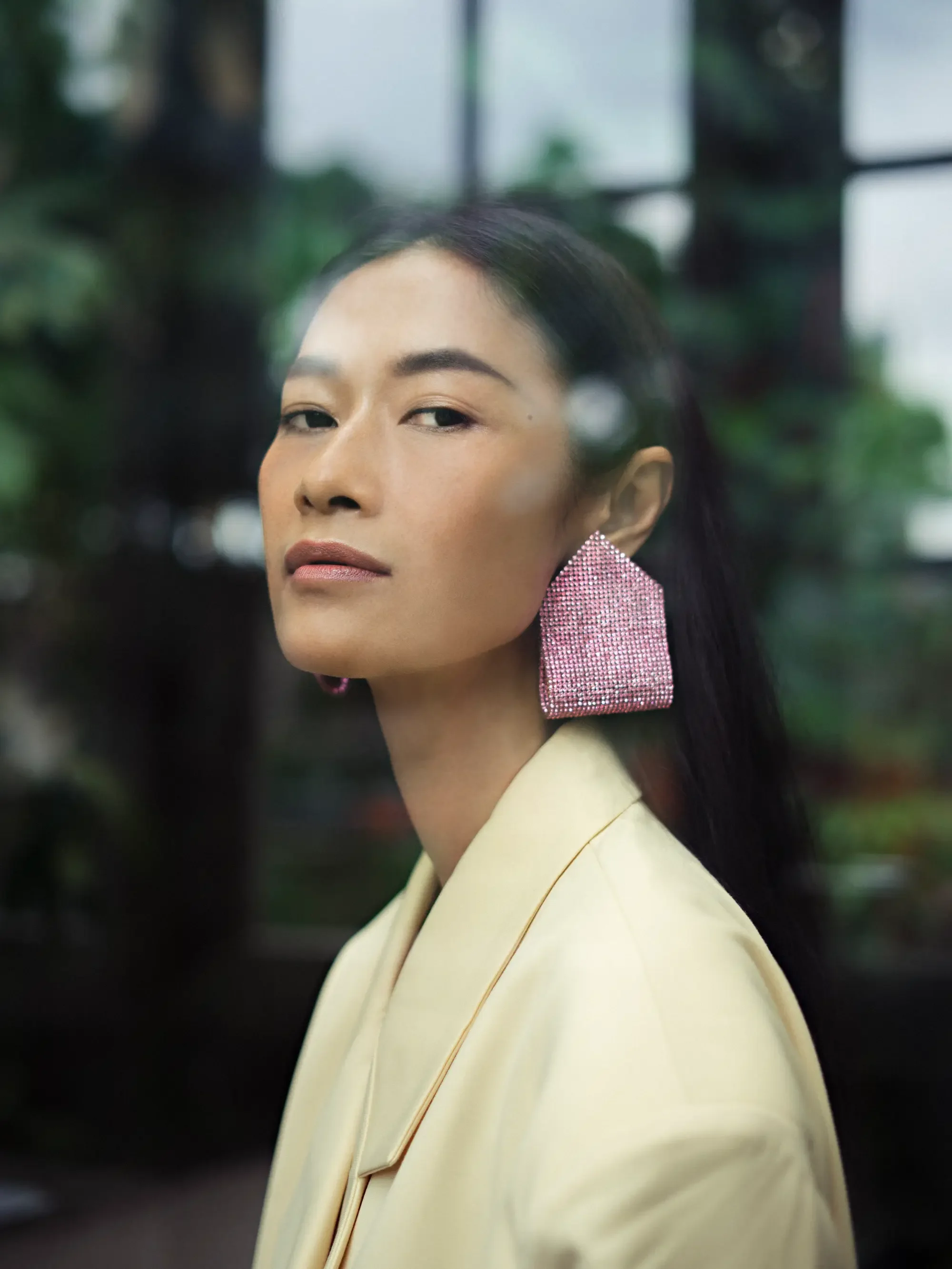 Close-up of a woman with dark hair, wearing a large pink, rhinestone-encrusted earring and a light-colored blazer, with a blurred background of greenery and windows.