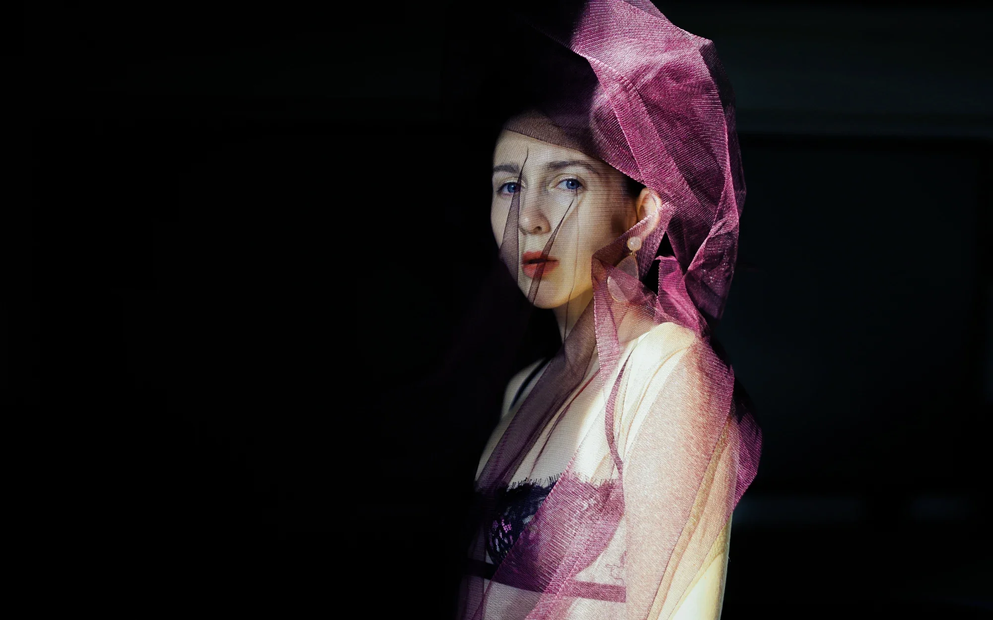 A woman dressed in traditional Japanese attire with a pink veil over her head, standing against a dark background.