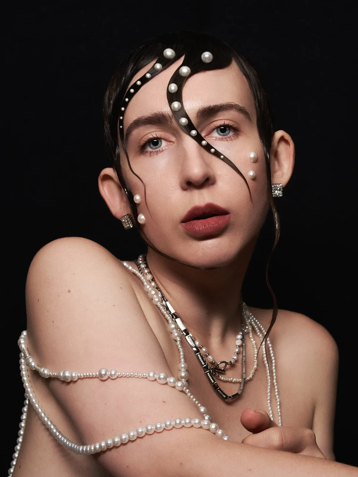 A woman with short wet dark hair adorned with pearl beads, wearing multiple pearl necklaces, earrings, and black and white jewelry, posing against a black background.