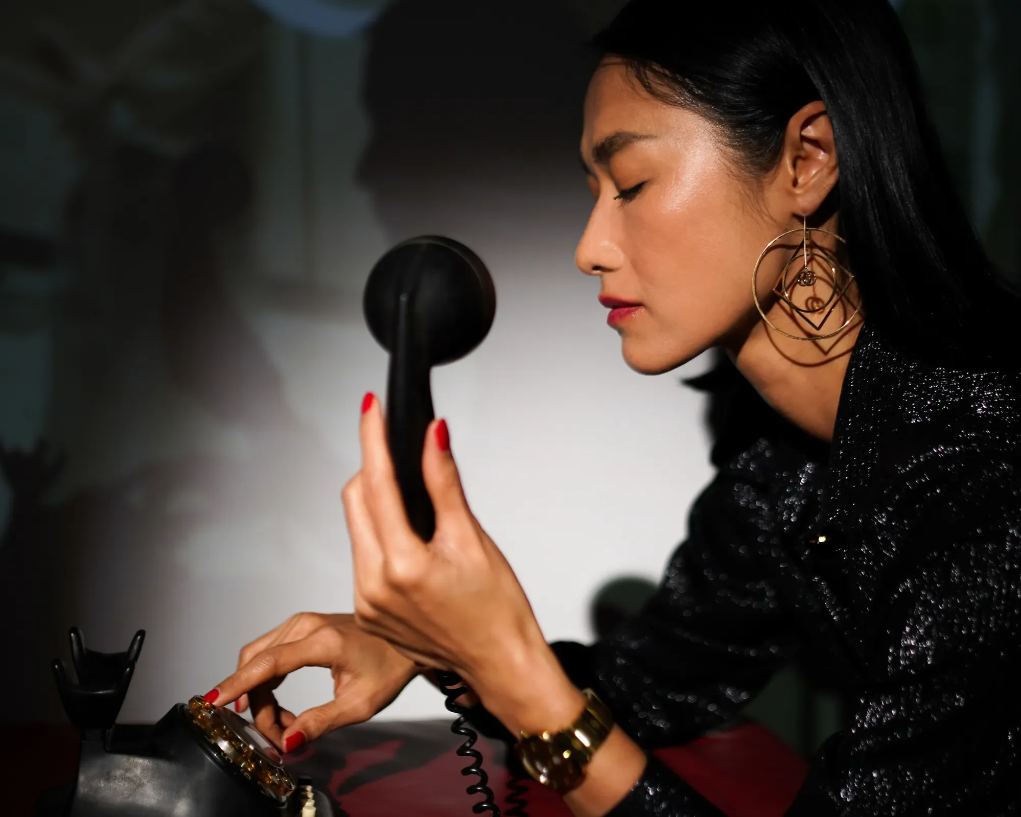 A woman with black hair, wearing large earrings, is looking down at an old-fashioned black telephone with a cord, holding the receiver in her right hand.