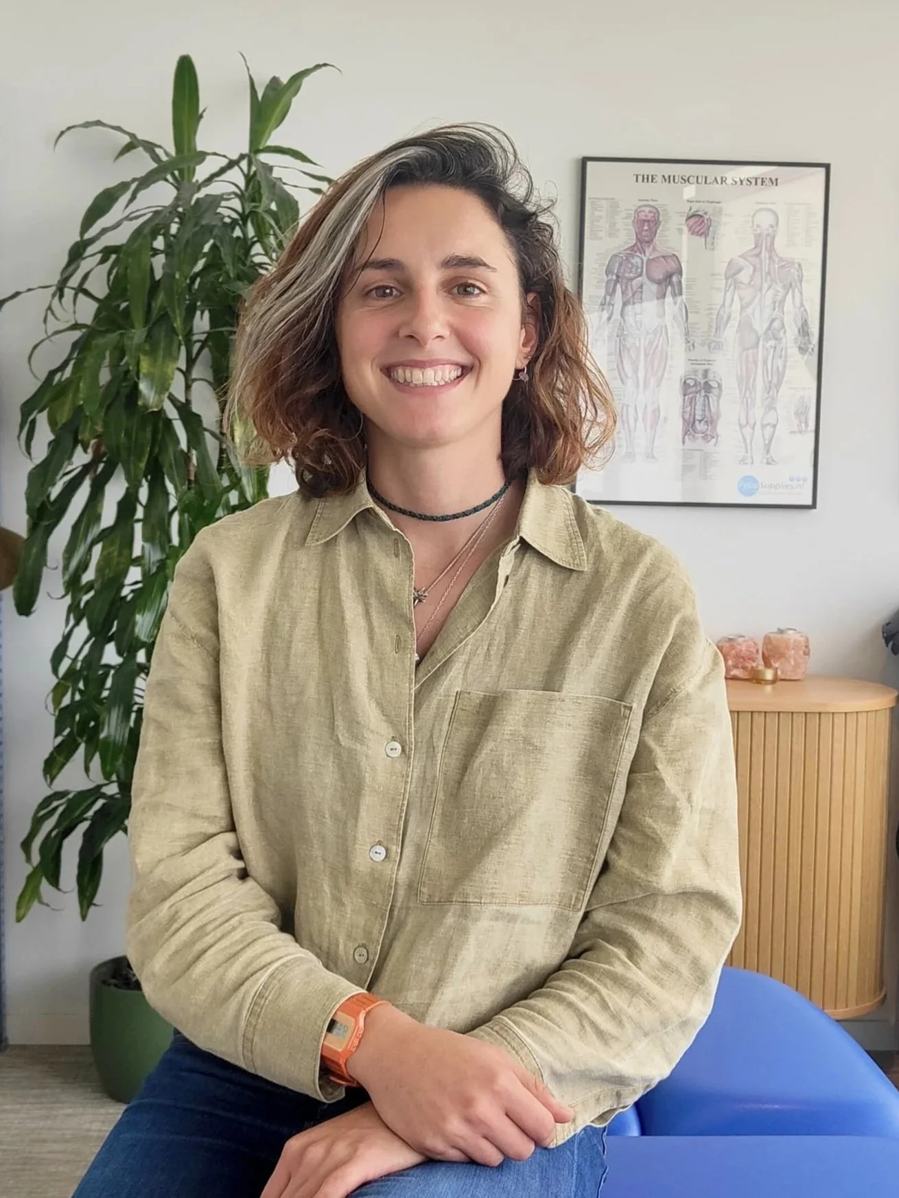 A woman with short wavy hair and a big smile sitting indoors near a large plant and a framed diagram of the muscular system in the background.