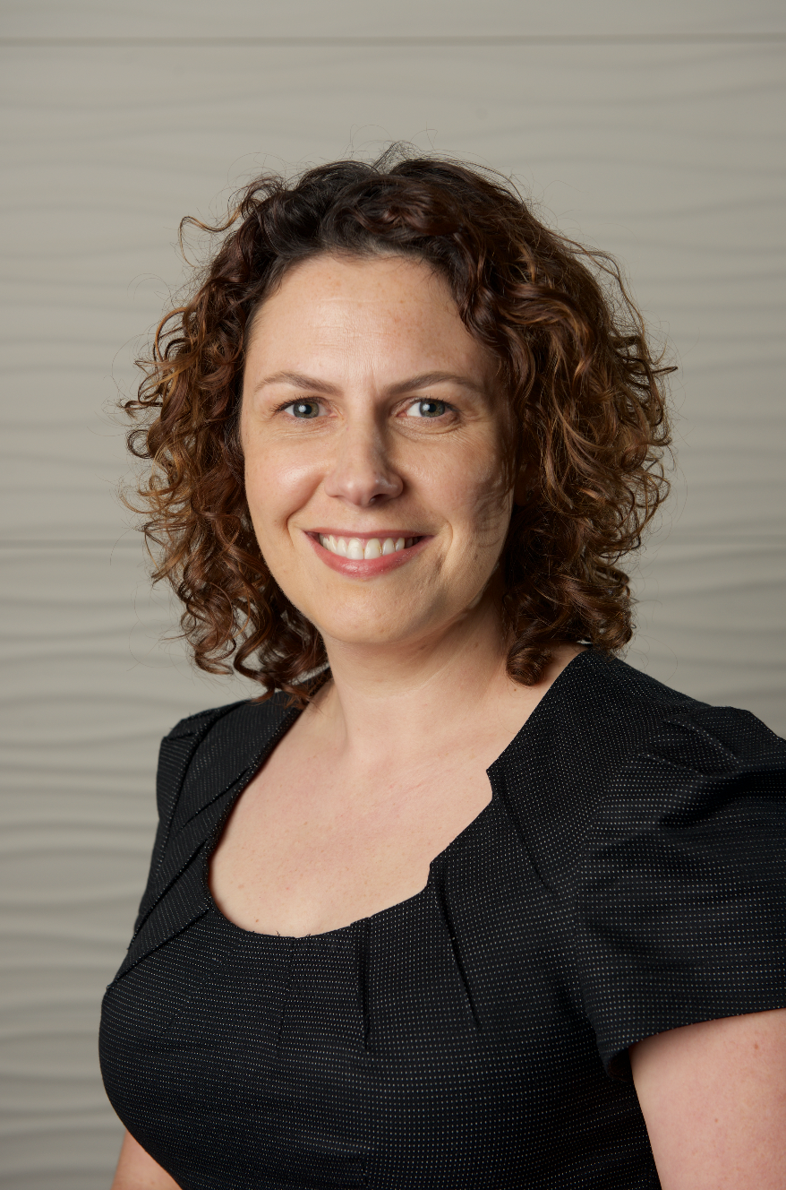 Jodie is smiling at the camera, wearing a dark textured top, against  a neutral background.