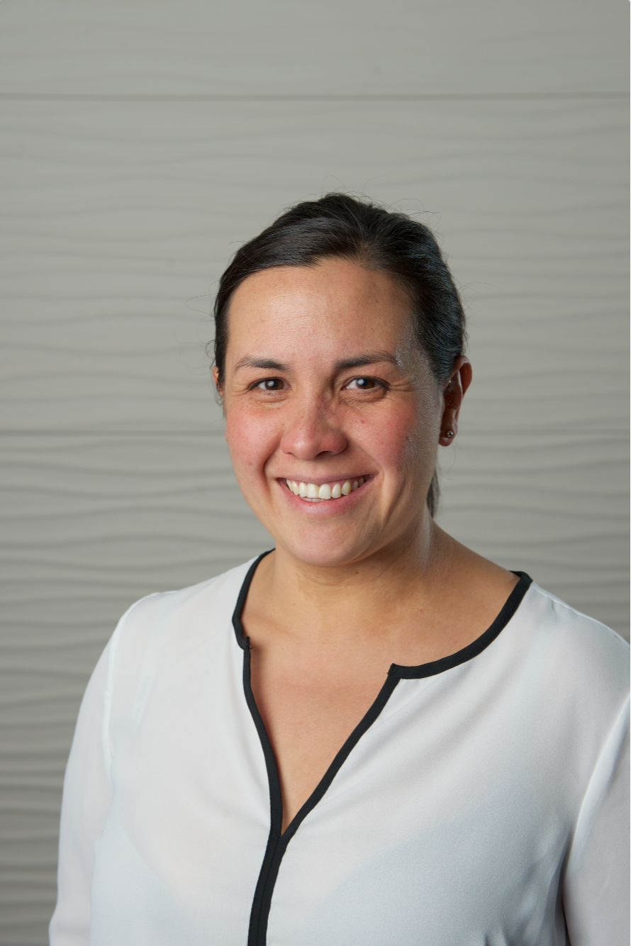 Emily is smiling at the camera, wearing a light coloured top, standing in front of a textured light grey background.