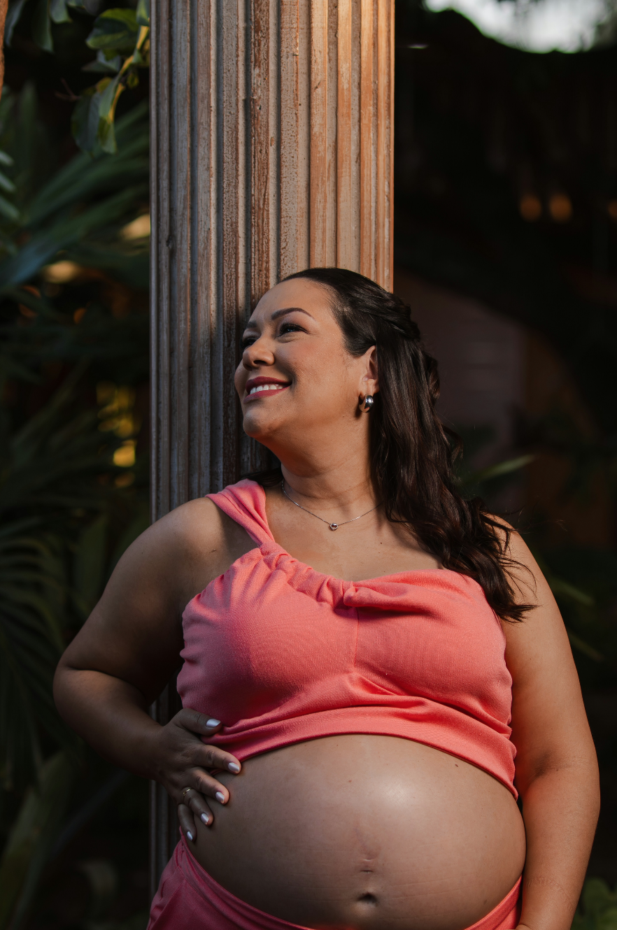 Pregnant woman smiling, wearing a sleeveless top, leaning against a wooden pillar outdoors with lush greenery in the background.