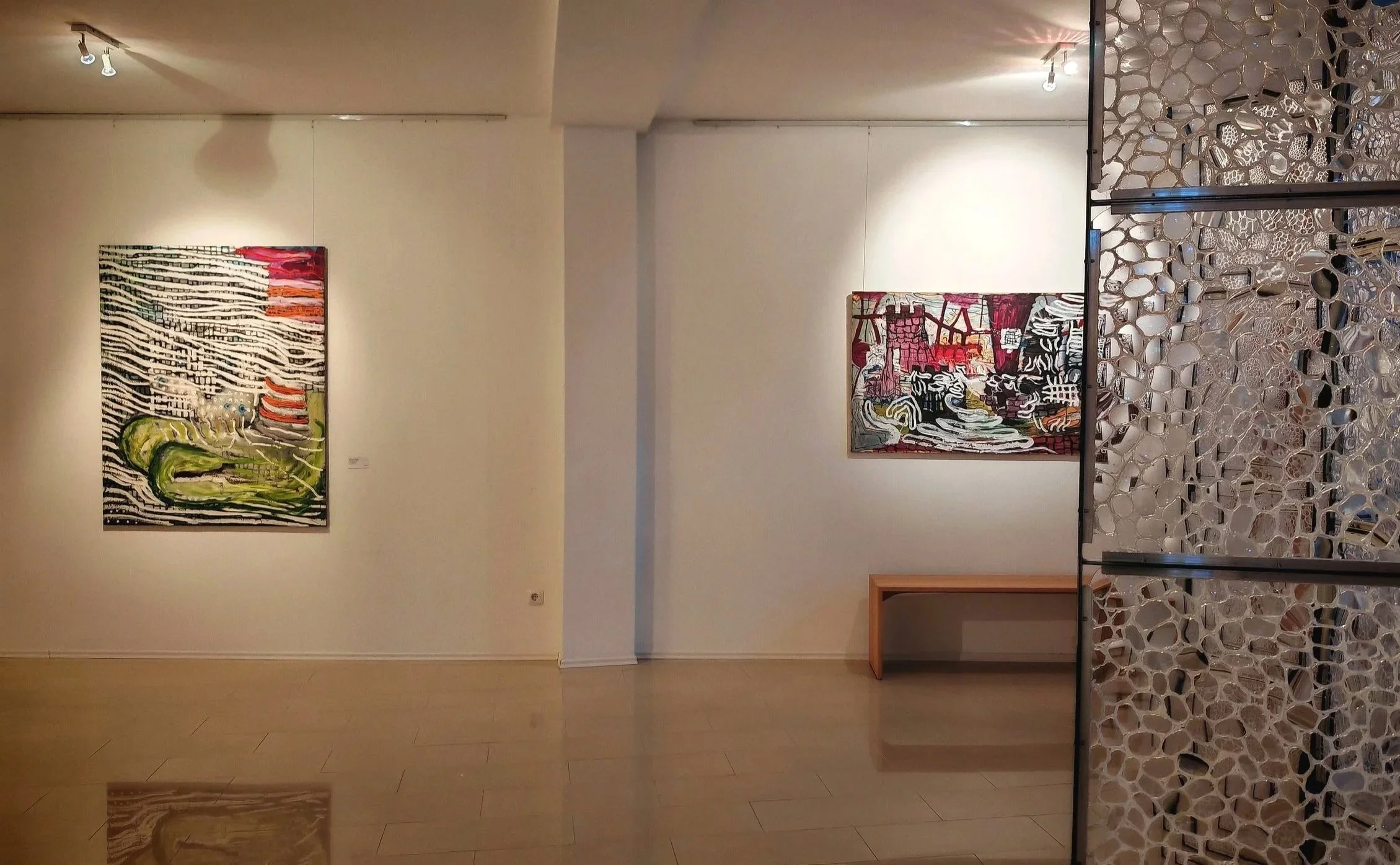 View of an art gallery with paintings on white walls, a textured partition on the right, and track lighting overhead.