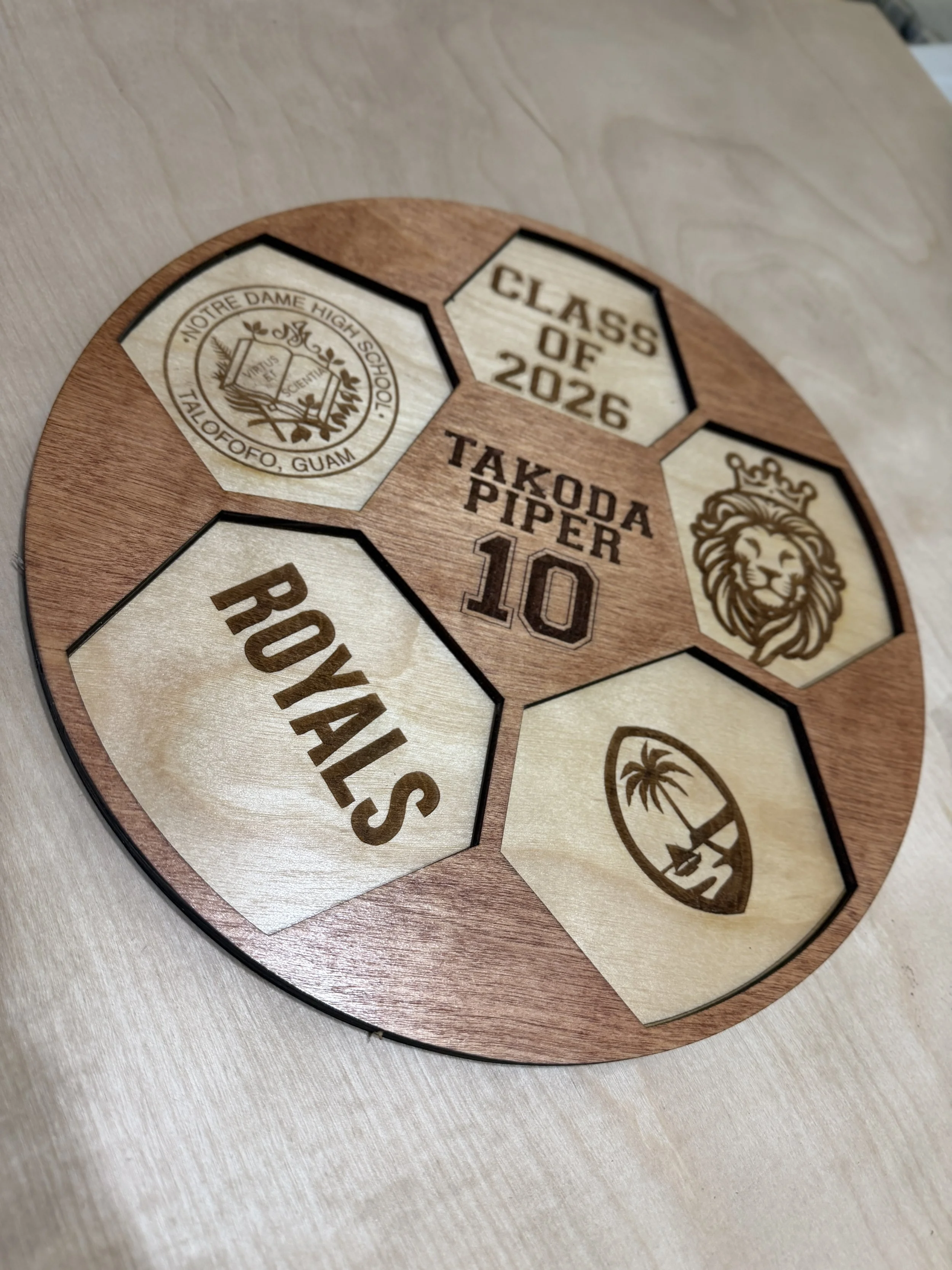 A wooden plaque with engraved names and logos for Notre Dame High School in Guam, Class of 2026, Takoda Piper, and others, including a lion and a palm tree.