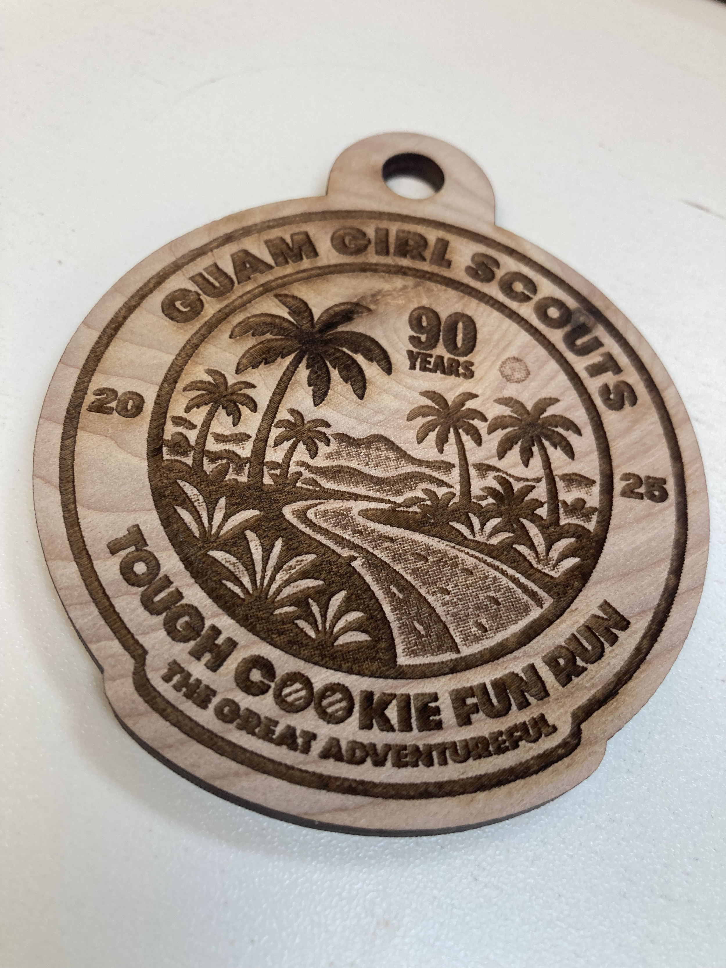 A circular wooden medallion engraved with a tropical scene of palm trees, mountains, and a winding river, with text indicating '90 years' of the CUAM Girl Scouts' outdoor adventure program.