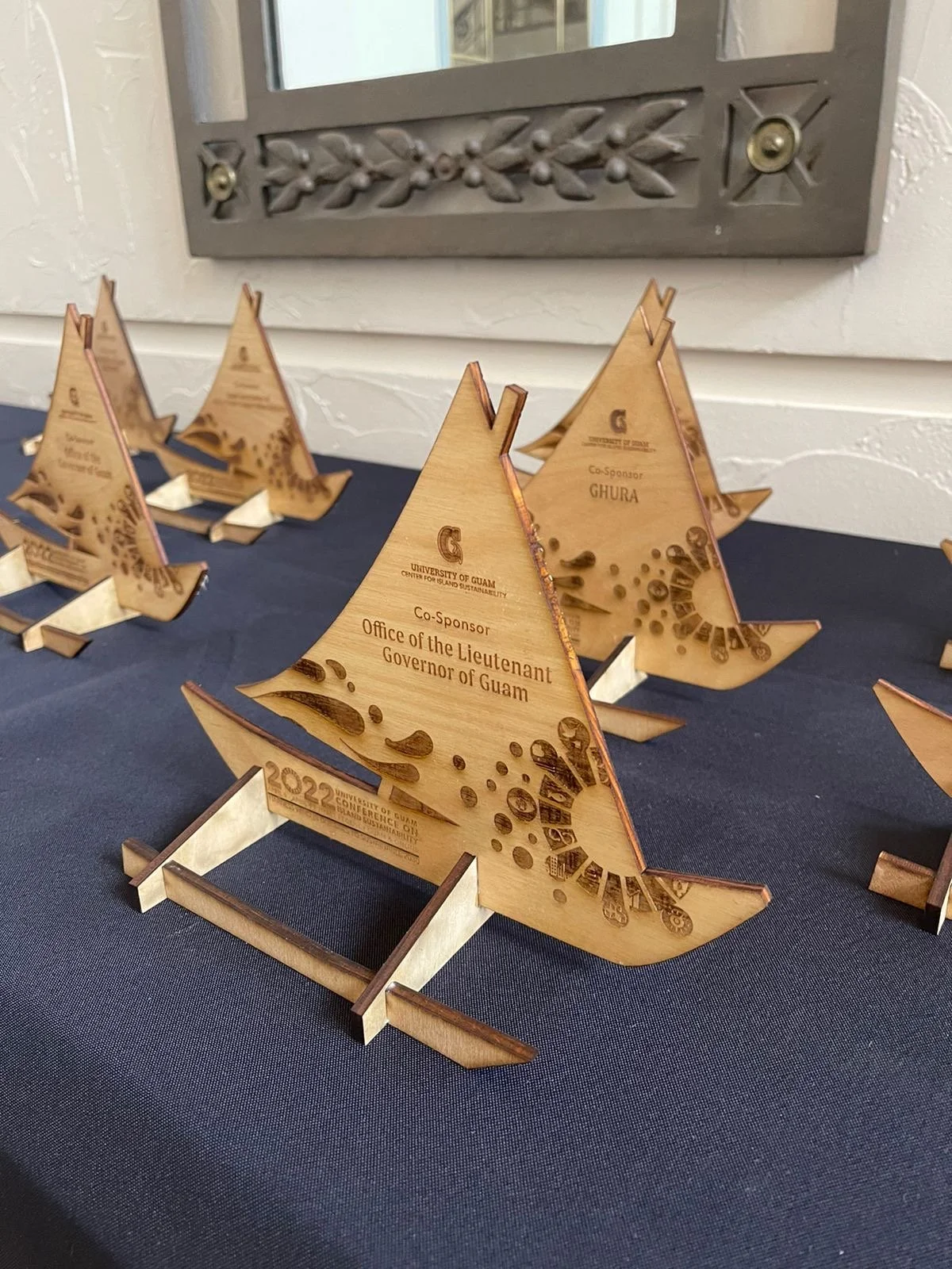 Wooden awards shaped like sailboats on a table, engraved with university and sponsorship details, with a mirror and textured wall in the background.