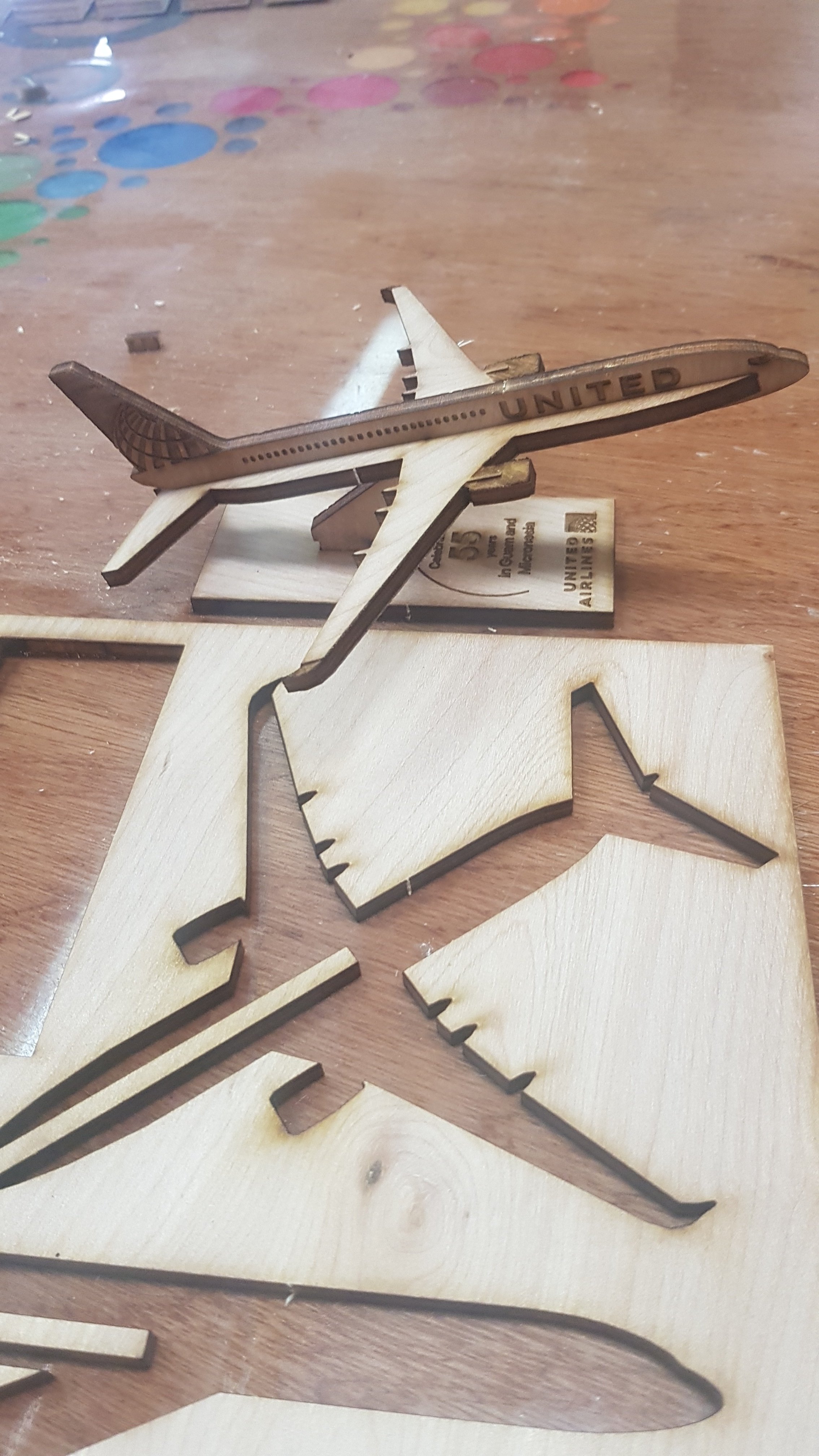 A wooden model airplane stating 'UNITED' on its fuselage, placed on a wooden table with colorful game pieces and cut-out wooden shapes in the background.