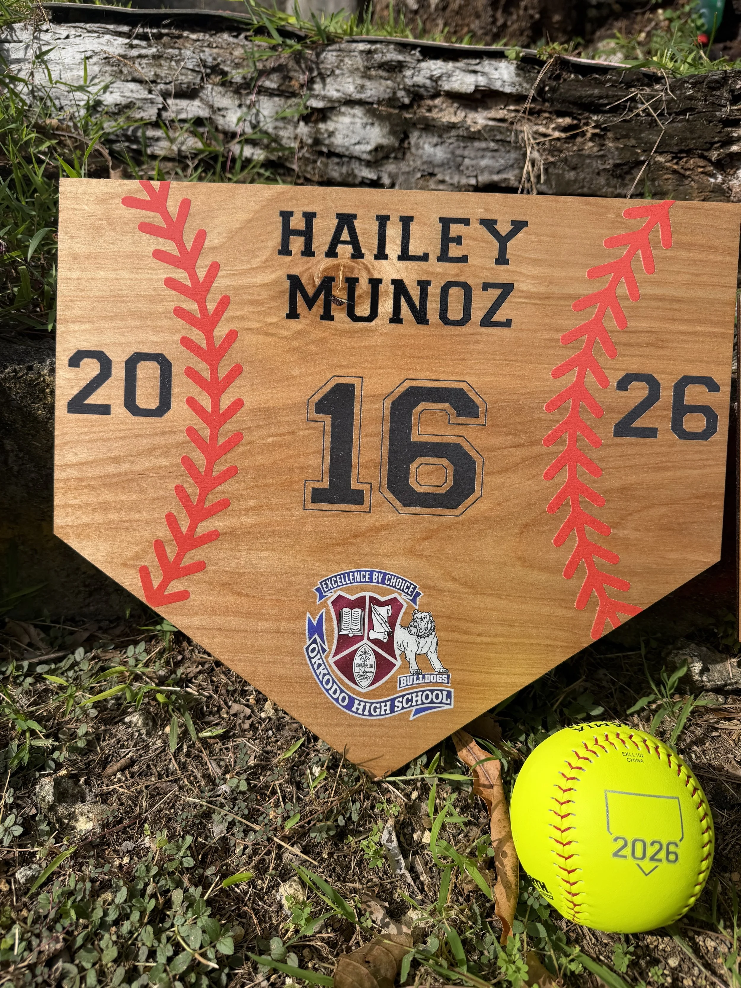 A baseball field tribute sign for Hailey Munoz with the year 2026, featuring a logo for Oxnard High School Bulldogs, placed on grass with soil and some leaves, and a bright yellow softball with red stitching and the year 2026 on it.