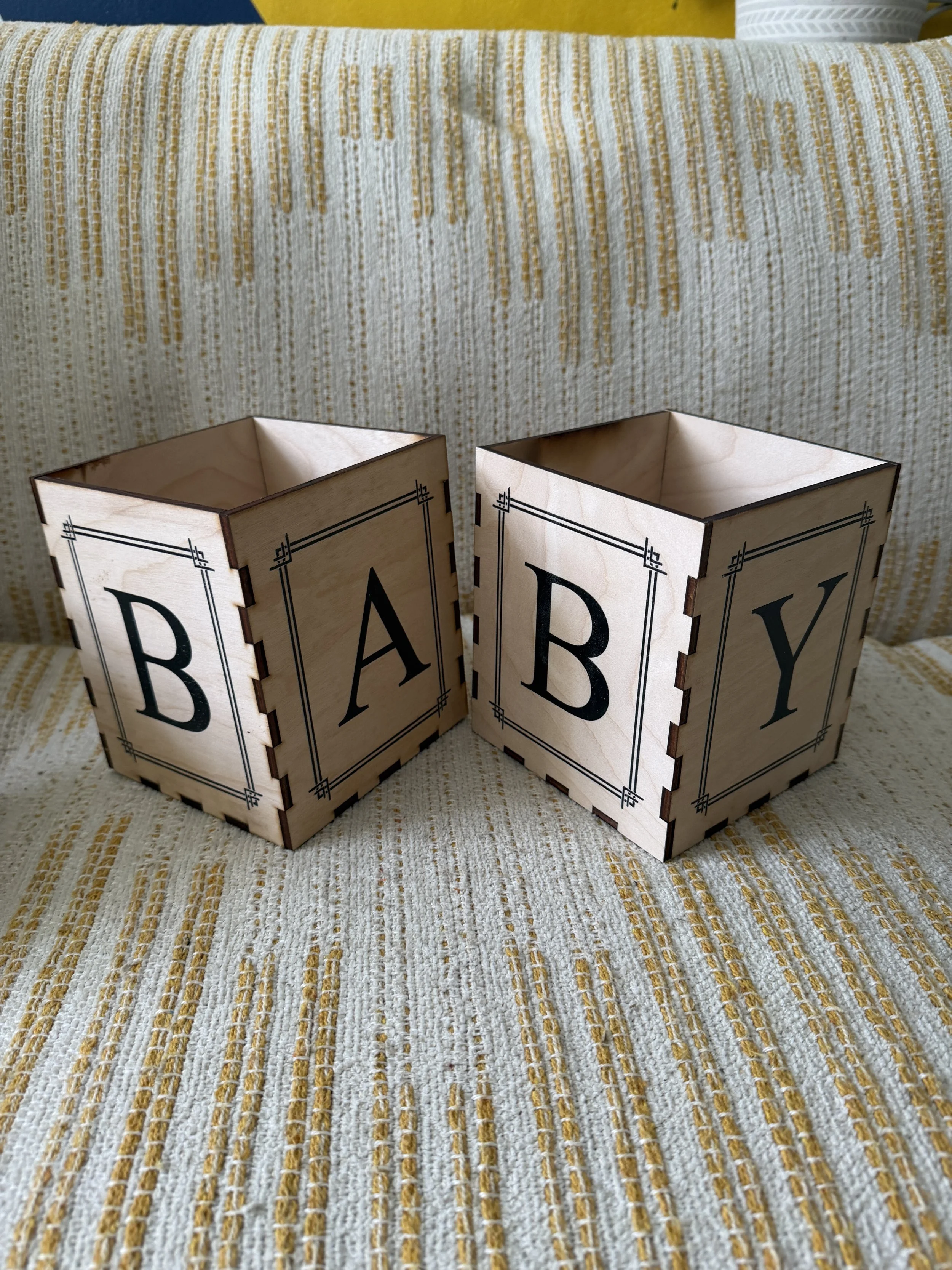 Two wooden boxes with the word "BABY" printed on their sides sit on a cream and yellow striped fabric surface.