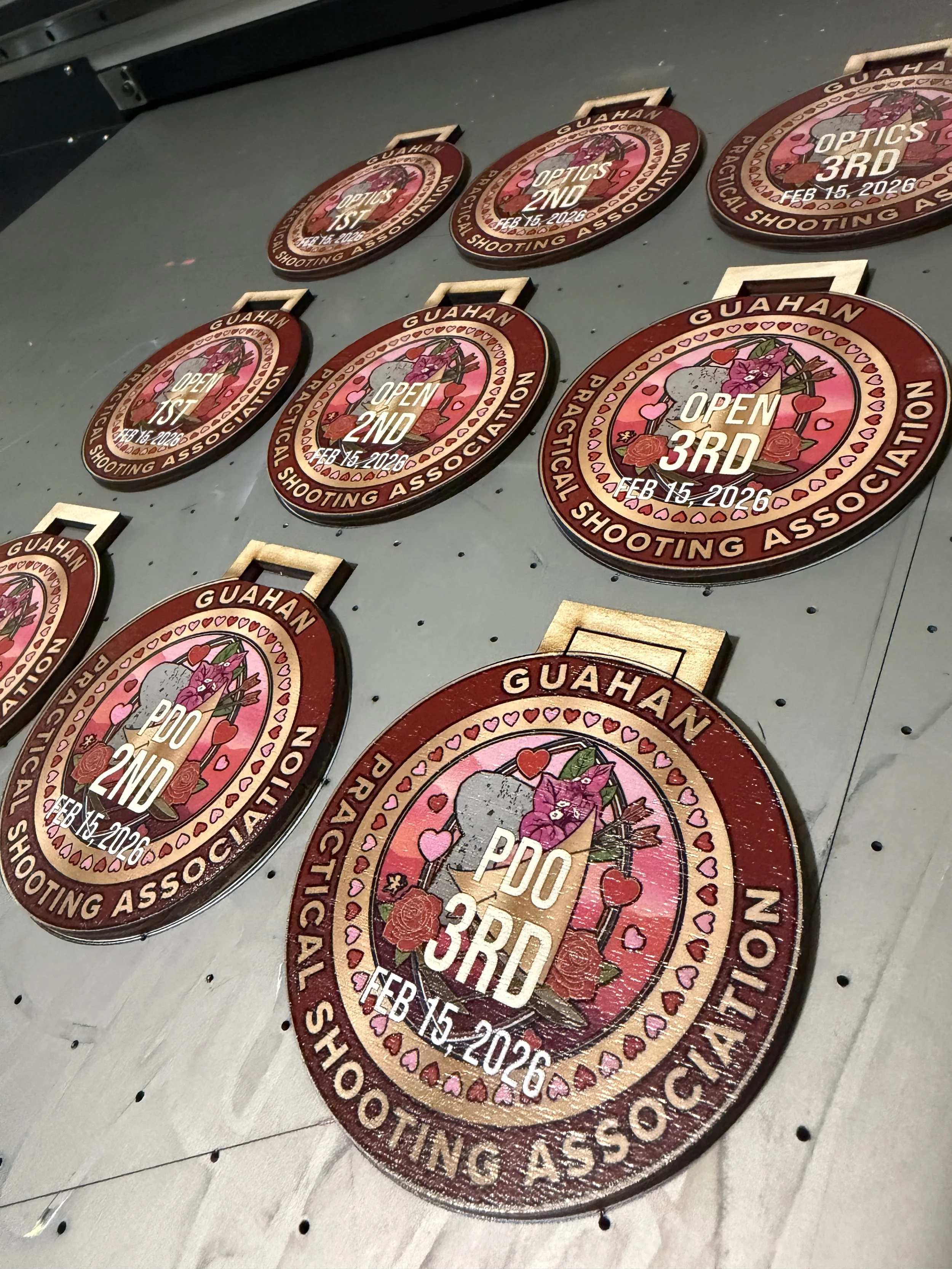 Medals from the Guahan Practical Shooting Association, indicating the winners of a shooting competition held on February 15, 2026. The medals are for first, second, and third place, with decorative pink hearts and roses surrounding the center text.