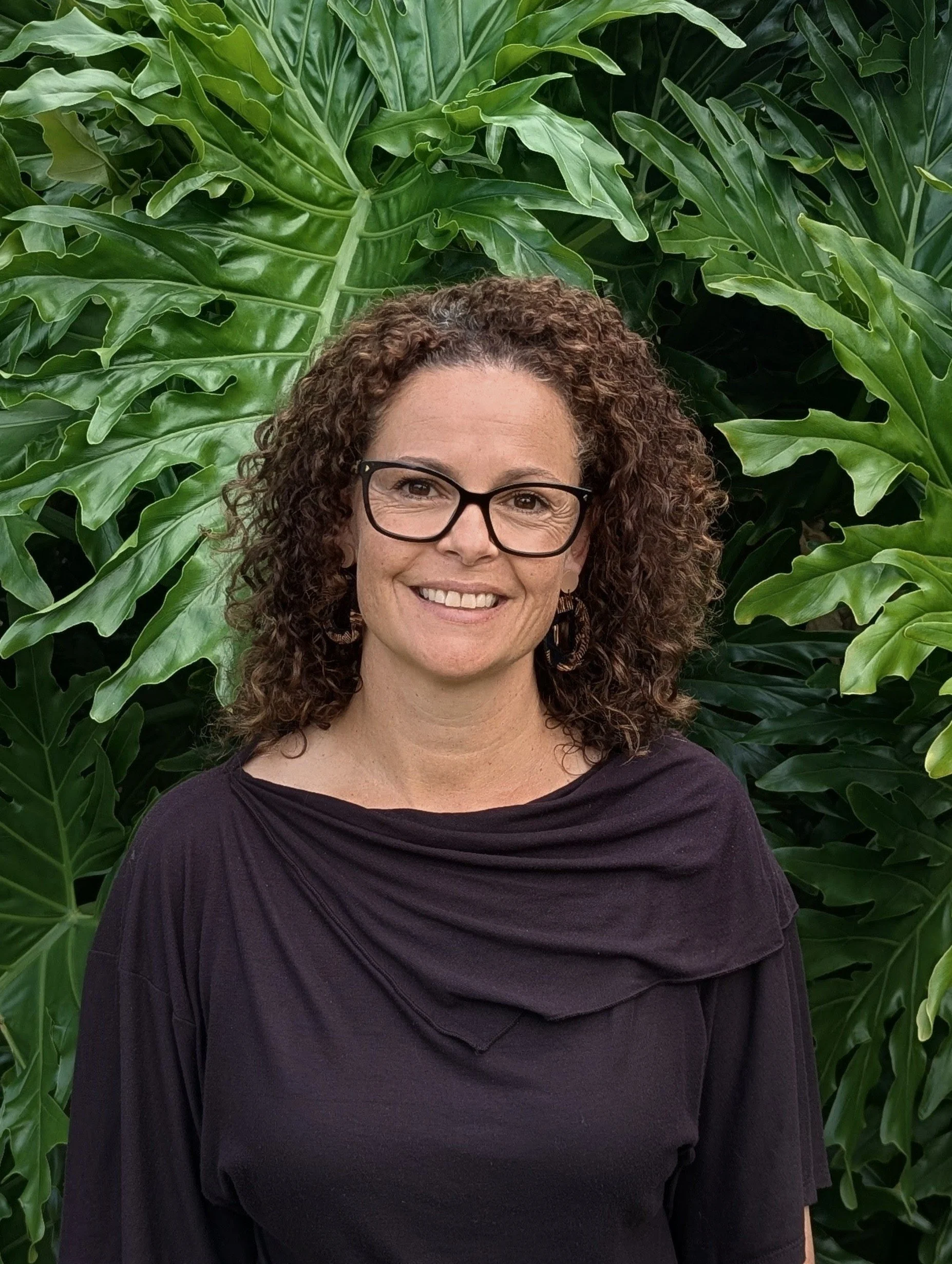 A woman with curly brown hair and glasses smiling, standing in front of large green tropical leaves.