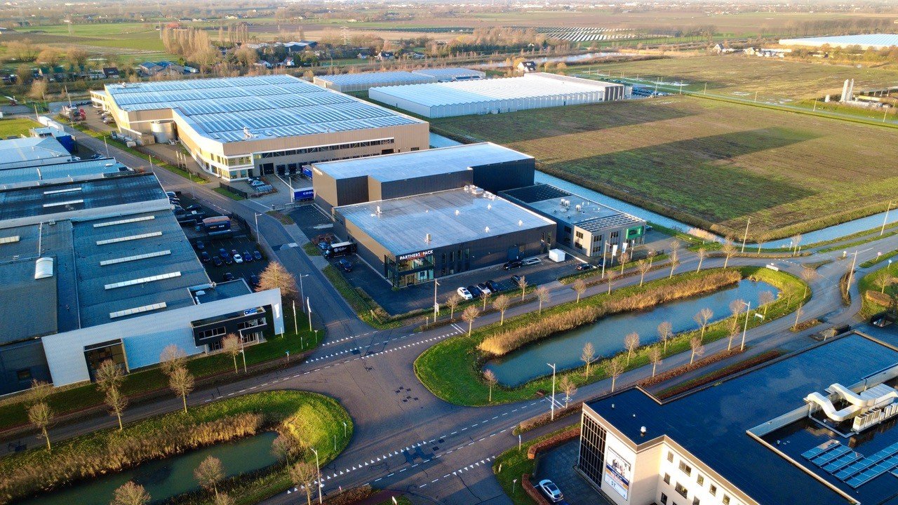 Een overzicht van een bedrijfsterrein met verschillende gebouwen, parkeerplaatsen, kleine waterpartijen en omliggende velden of landbouwgrond.