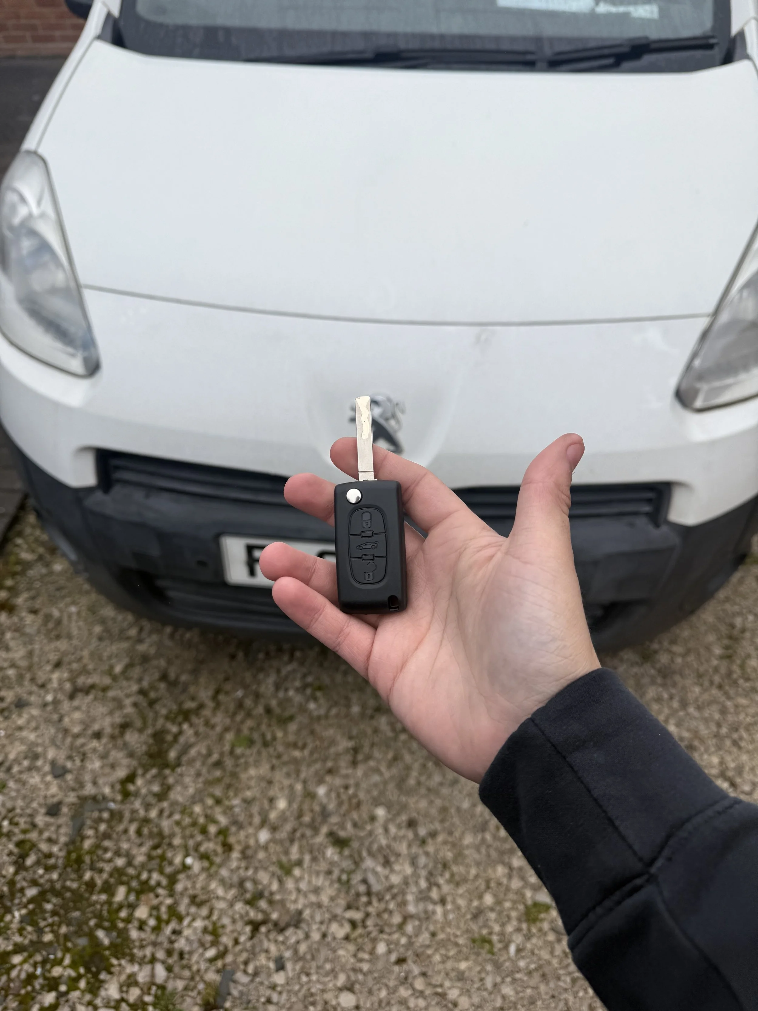 A person holding a car key in front of a white car, viewed from above, with gravel ground beneath.