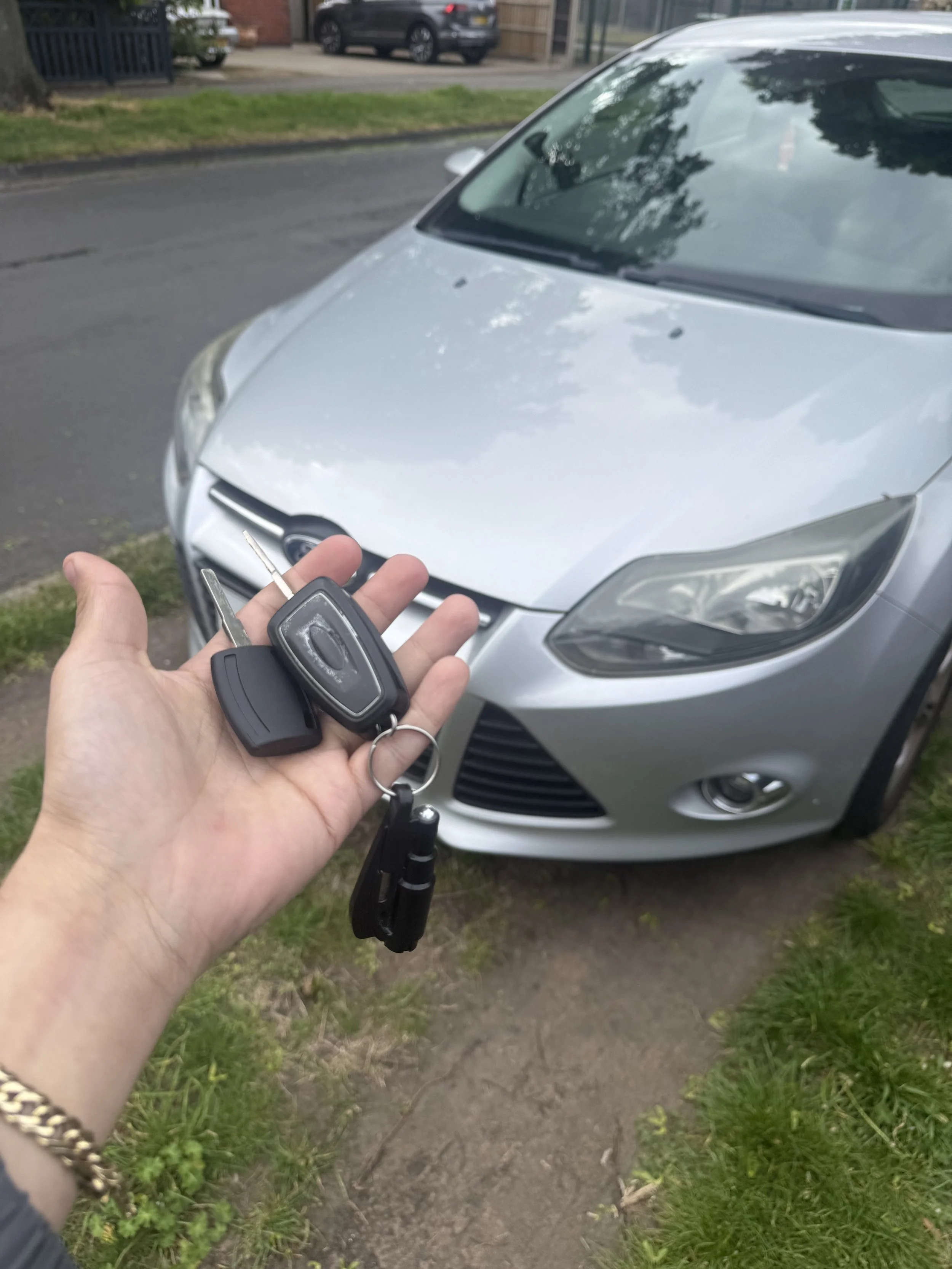 A person holding car keys in front of a silver car parked on a grassy area next to a sidewalk.