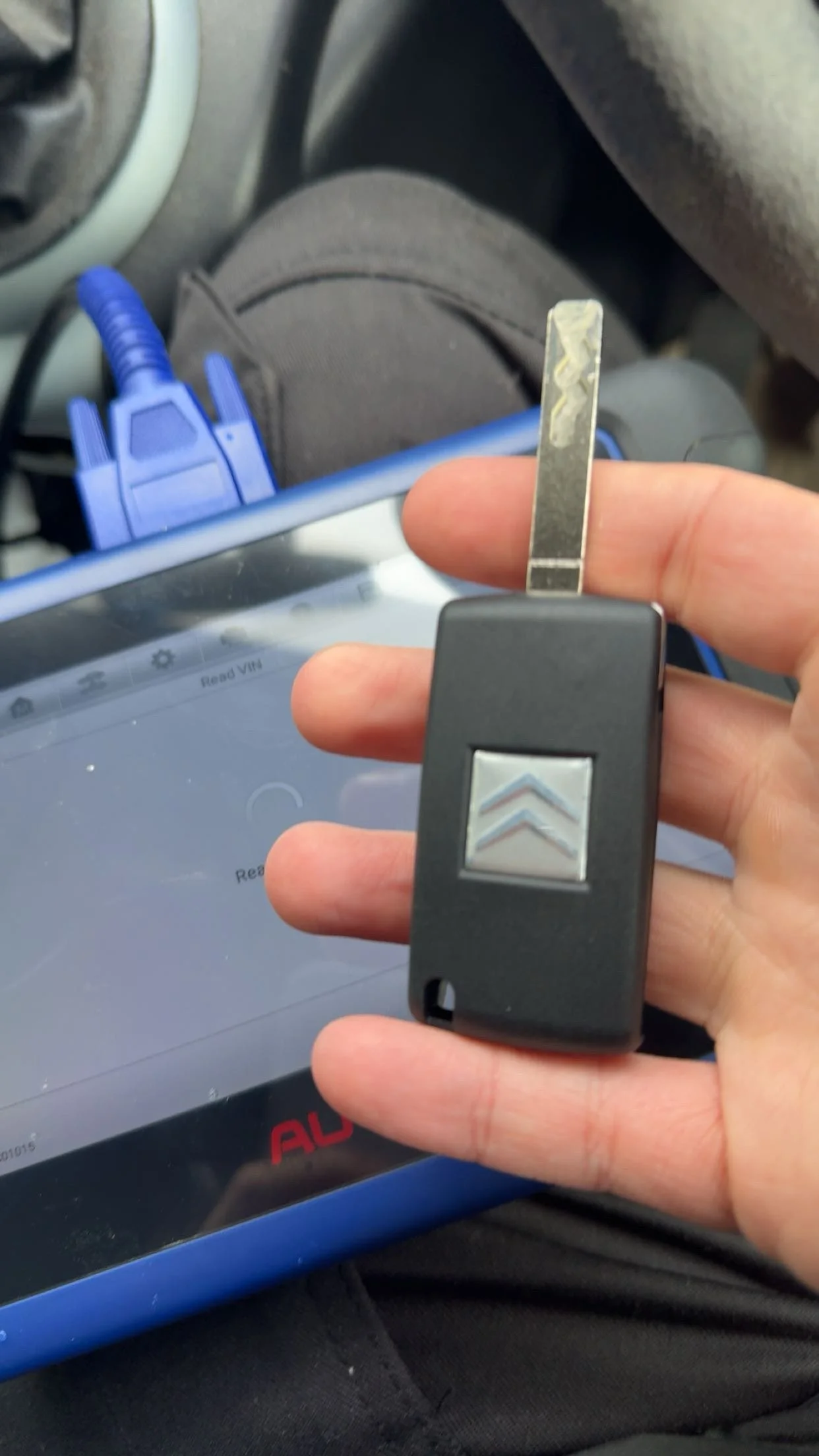 Close-up of a hand holding a car key with a metallic chip, in front of a tablet device displaying an app, with a car interior and a blue cable in the background.