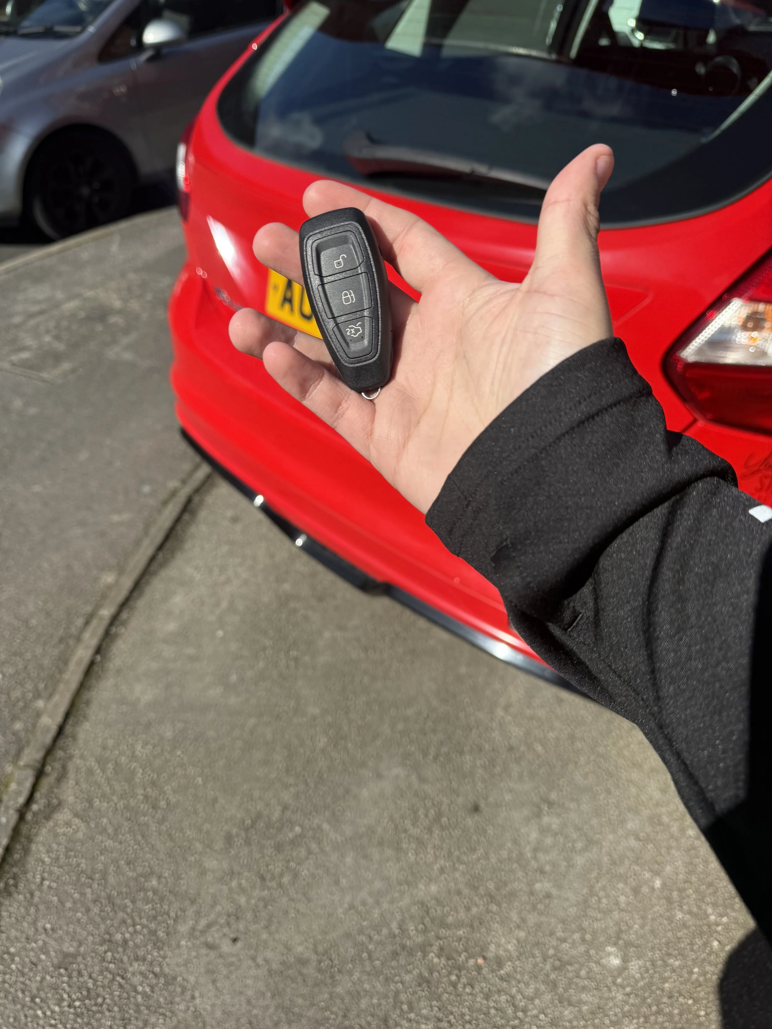 A hand holding a car key fob in front of a red car parked on the street.