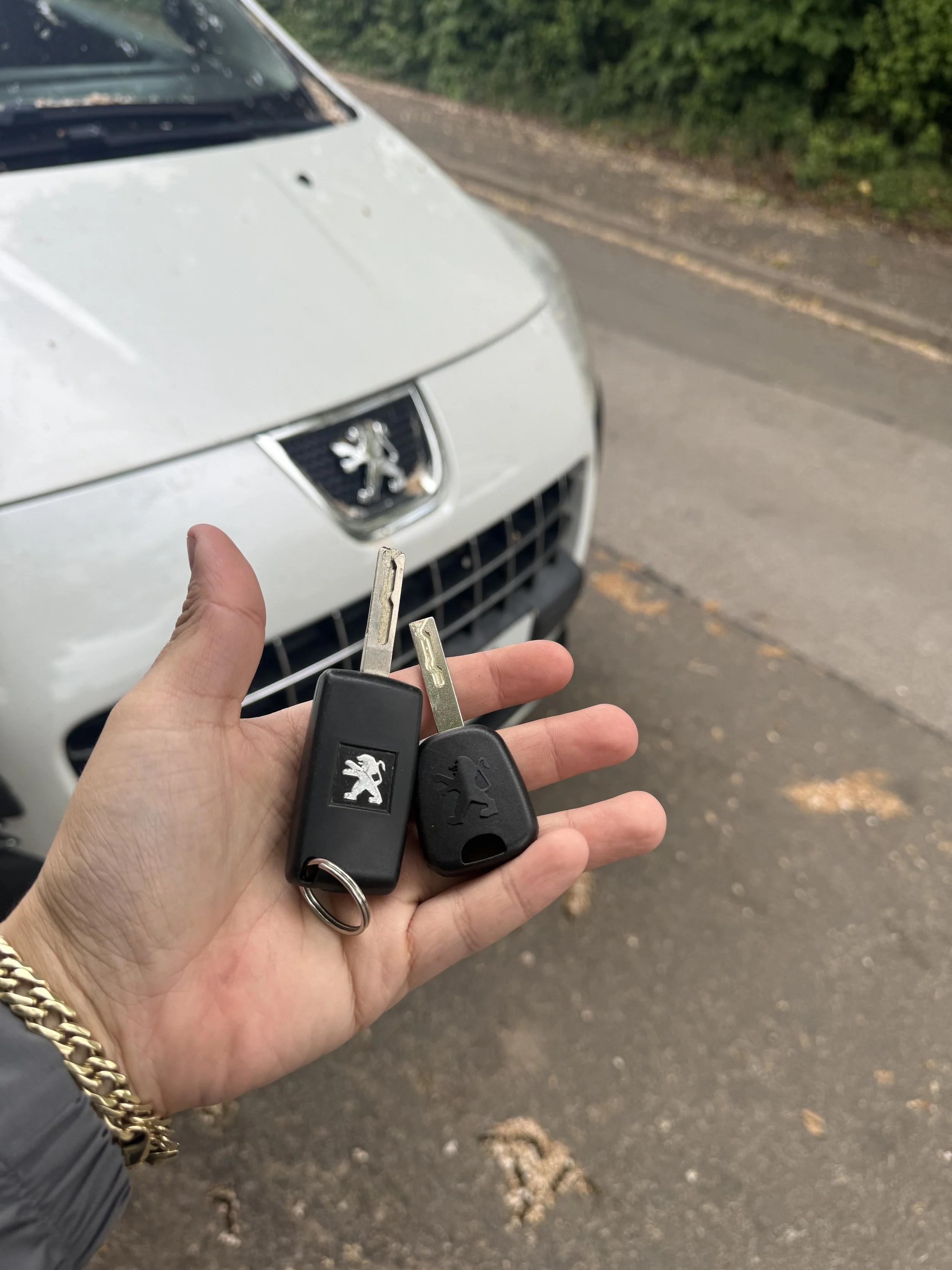 A hand holding car keys in front of a white Peugeot car on a road with green trees in the background.