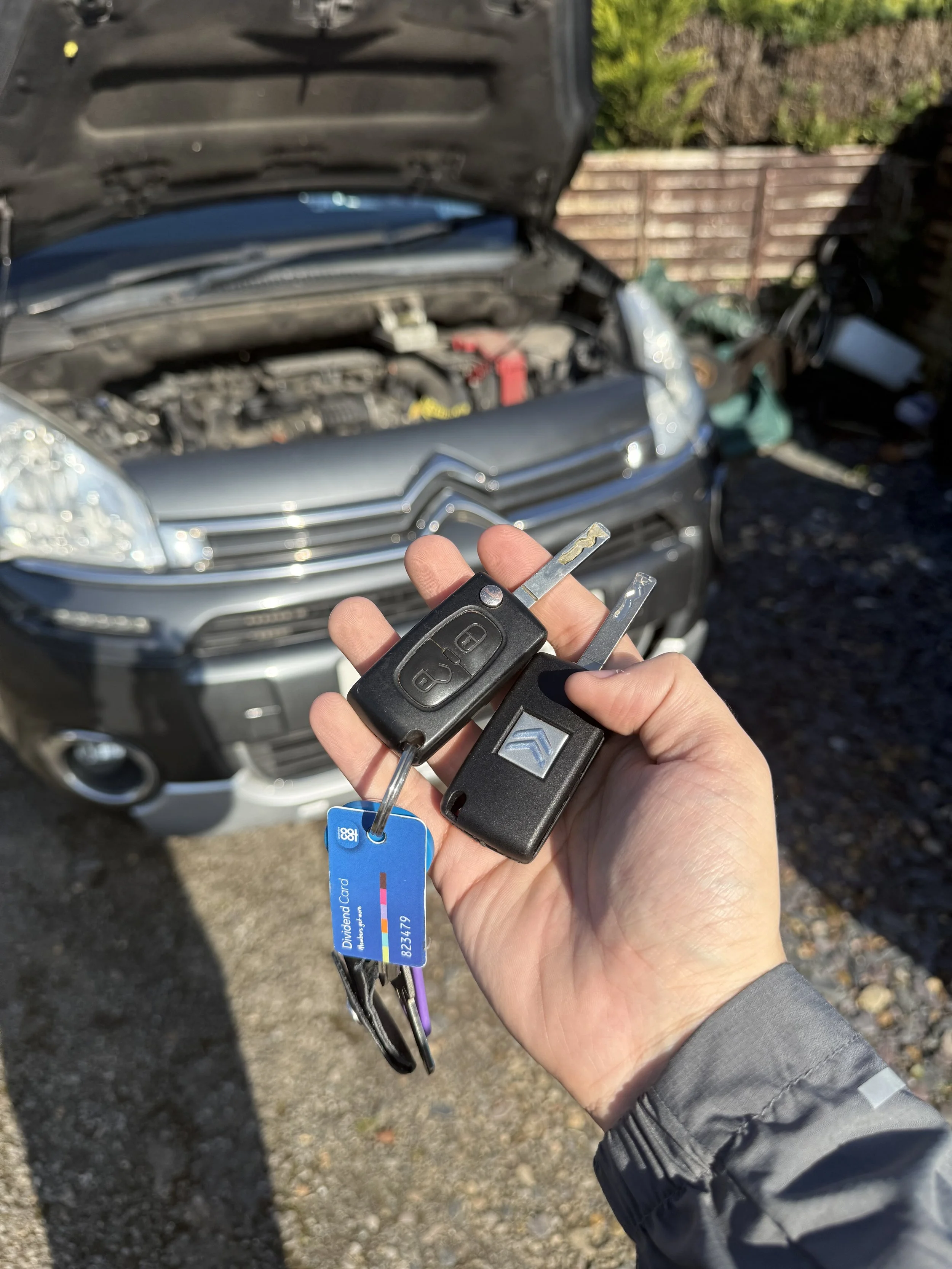 A person holding car keys and a bank card in front of an open car hood, with a blurred view of the car's engine and a wooden fence in the background.