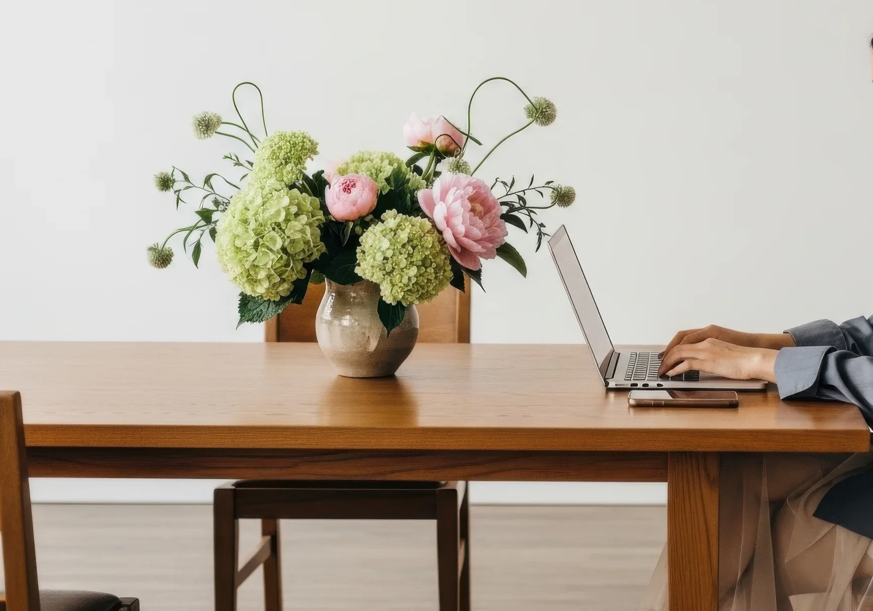Eine Blume mit Rosatönen und grünlichen Hortensien in einer Keramikvase auf einem Holztisch, daneben eine Person, die an einem Laptop arbeitet.