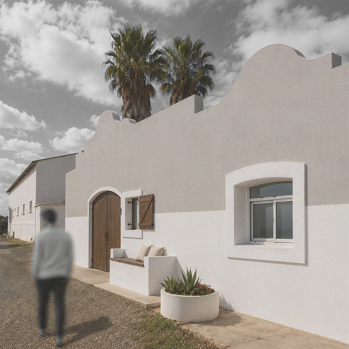 Une maison blanche au style méditerranéen avec un mur blanc, une fenêtre à volet en bois, un banc avec des coussins, un pot avec des plantes succulentes, et deux palmiers en arrière-plan sous un ciel partiellement nuageux.