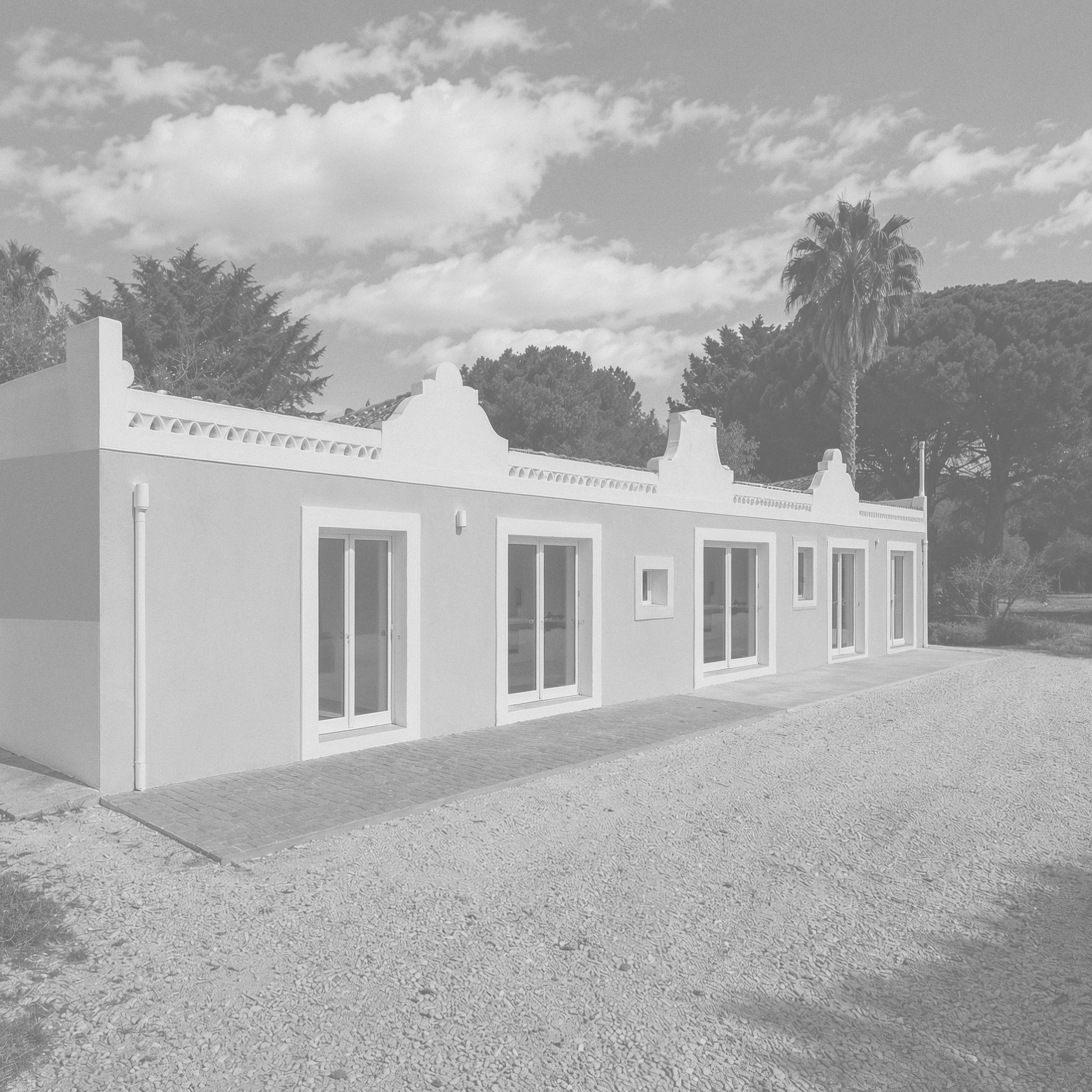 Vue d'une maison blanche avec toit de tuiles, entourée d'arbres dont un palmier, sous un ciel avec quelques nuages.
